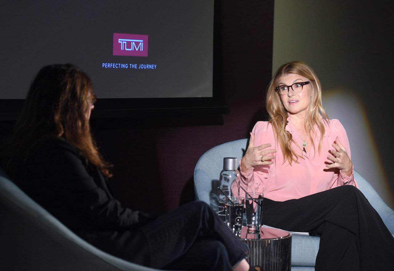 A spotlight shines on Connie Britton, seated and wearing glasses, a pink top, and black pants