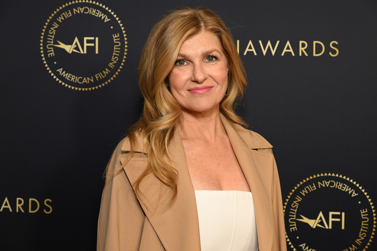 Connie Britton in a white top and brown jacket with her hair down, posing and smiling