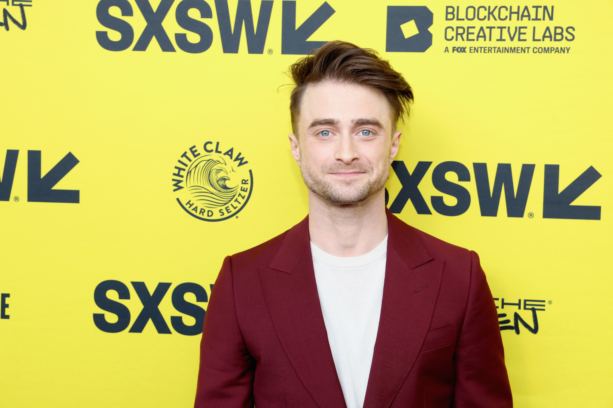 Daniel Radcliffe on the red carpet at SXSW. He's standing in front of a bright yellow wall and wearing a red jacket and white shirt.