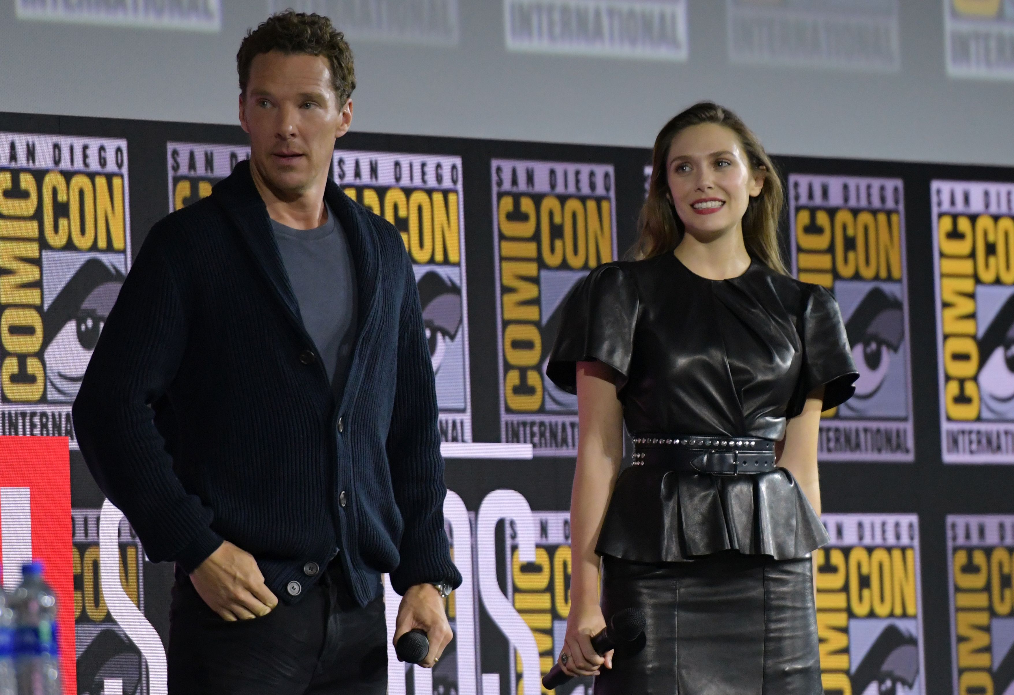 Benedict Cumberbatch and Elizabeth Olsen, who star as Doctor Strange and Scarlet Witch in 'Doctor Strange 2,' stand onstage during a panel. Cumberbatch wears a dark blue cardigan over a blue shirt and black pants. Olsen wears a black leather shirt over a black leather skirt.