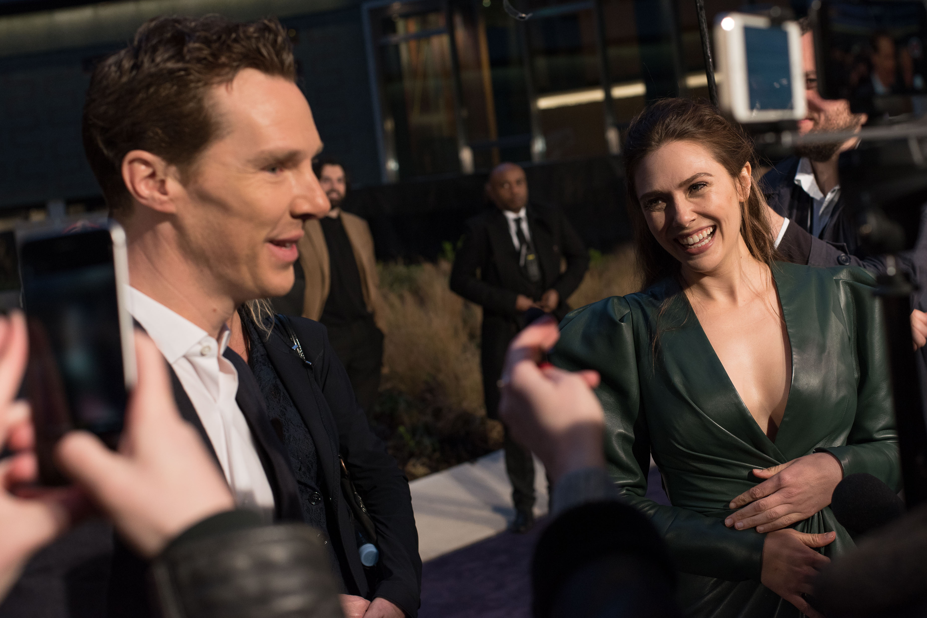 Benedict Cumberbatch and Elizabeth Olsen, stars of 'Doctor Strange 2' in the MCU, talk to press on the red carpet. Cumberbatch wears a black suit over a white button-up shirt. Olsen wears a dark green long-sleeved leather dress.