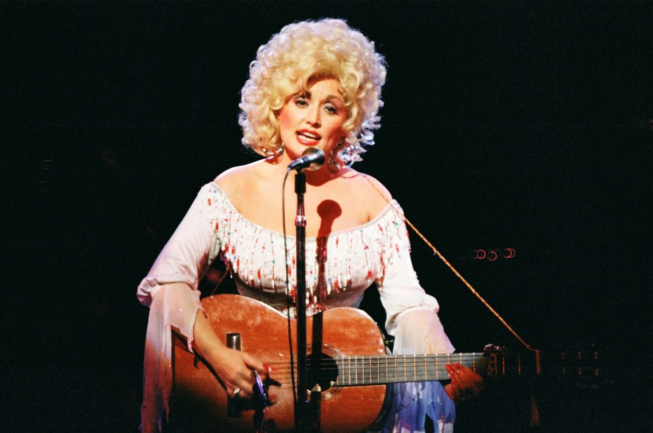 Dolly Parton wears a white dress and holds a guitar. She stands in front of a microphone.