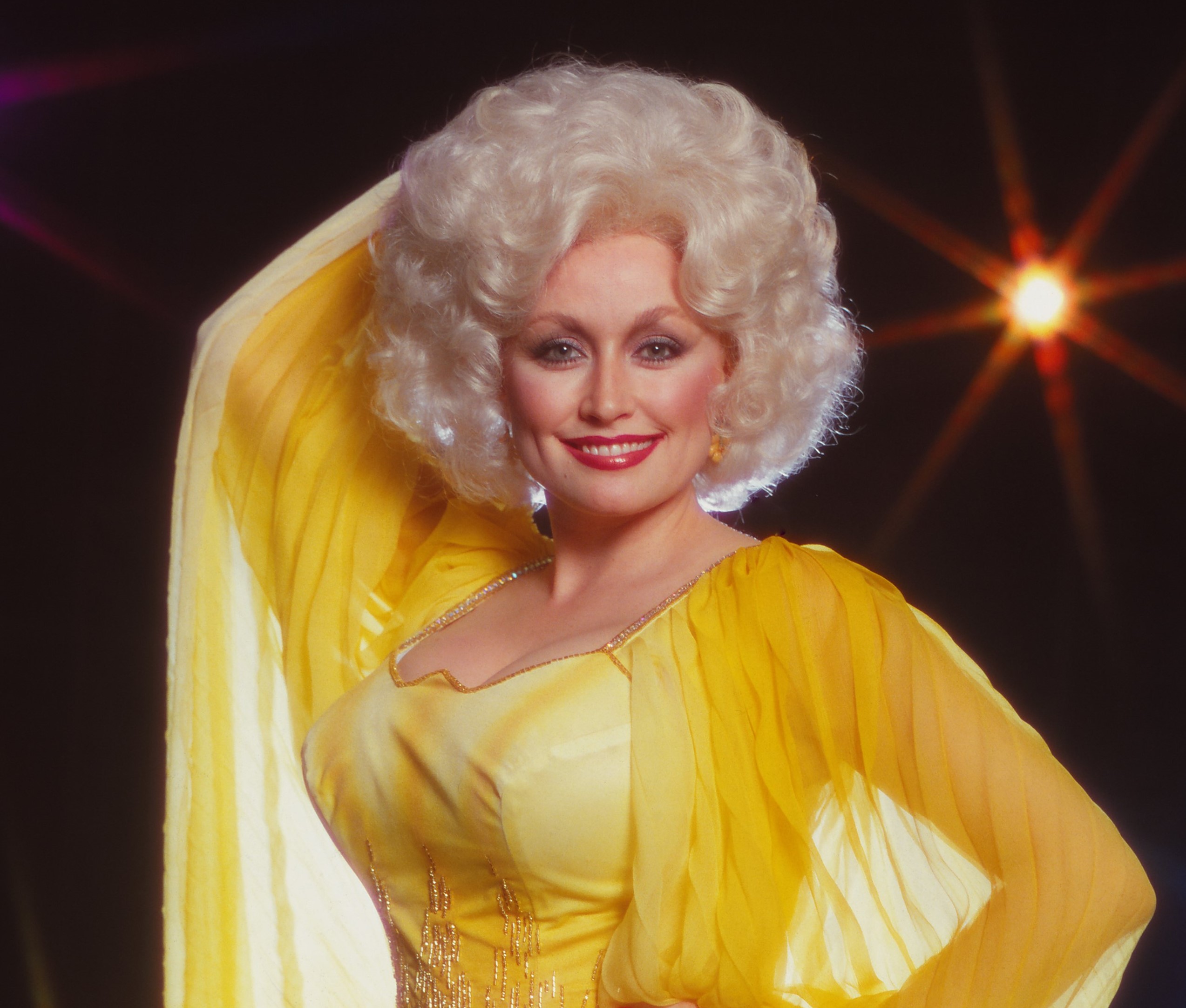 Dolly Parton wears a yellow dress and stands with one hand on her hip and one behind her head.