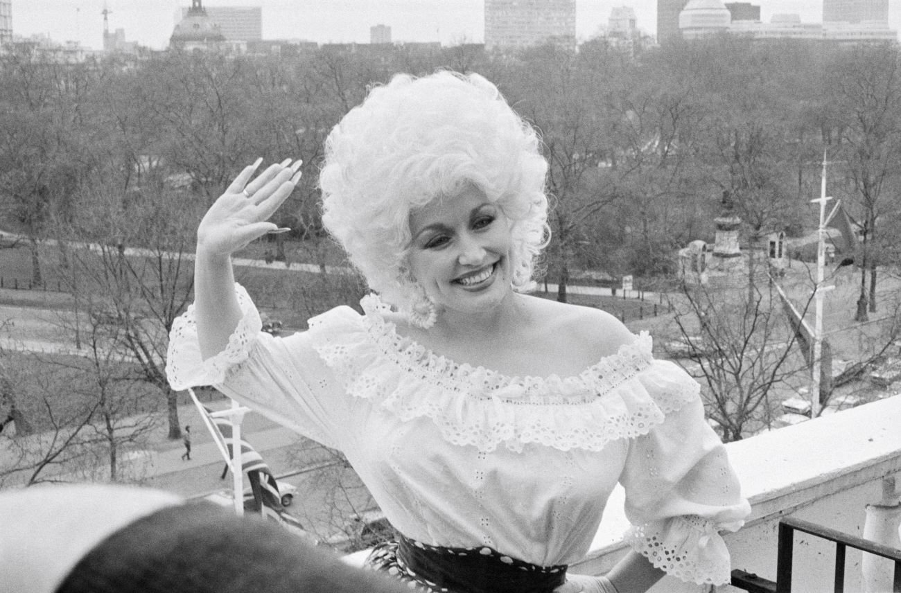 A black and white photo of Dolly Parton standing on a balcony and waving her hand.