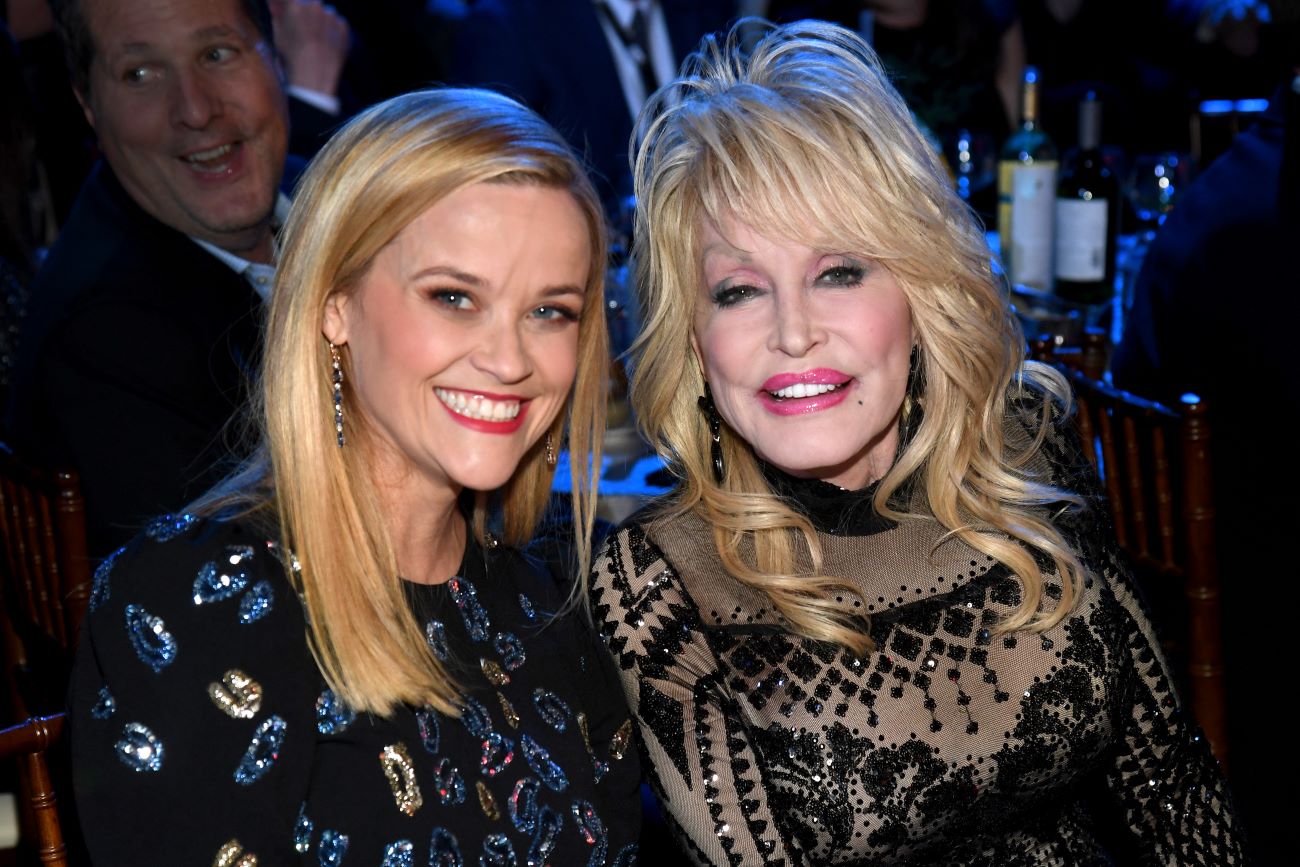 Reese Witherspoon and Dolly Parton sit next to each other and smile in a dark room.