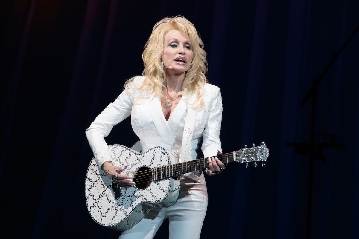 Dolly Parton wears a white outfit and plays a white guitar on stage.