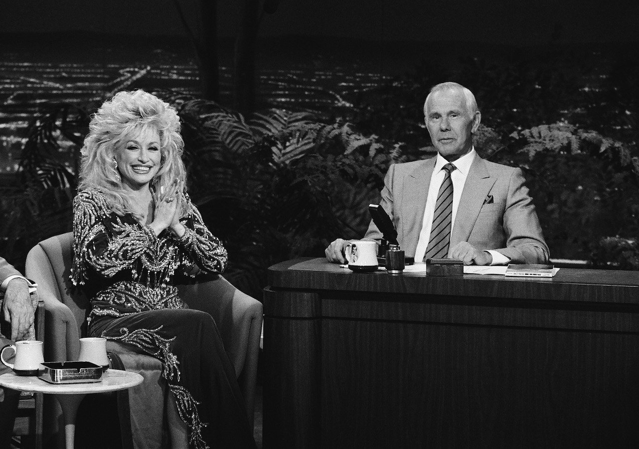 Black and white photo: Dolly Parton sits with her hands held in prayer form next to Johnny Carson on 'The Tonight Show' in 1990