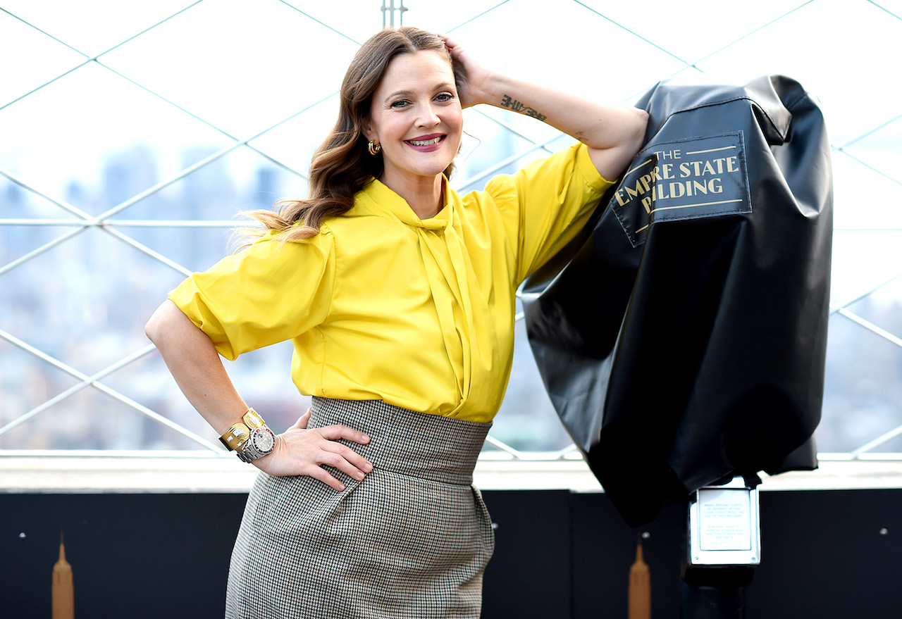 Drew Barrymore in a yellow top, smiling and leaned on one elbow with the other hand on her hip