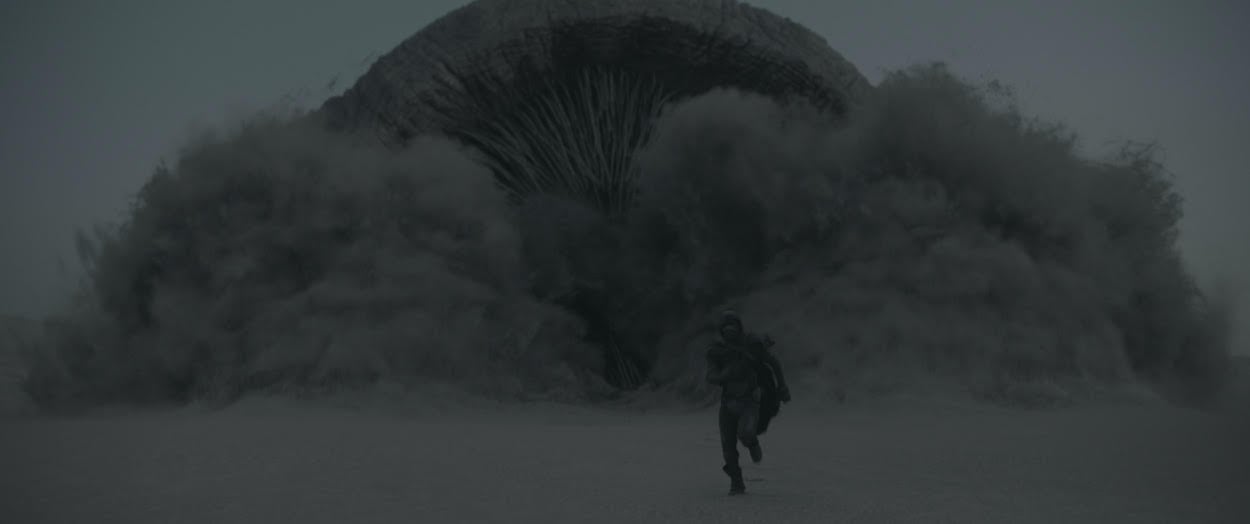 A sandworm in Dune opens its mouth as it breaches the sand while Timothee Chalamet's character, Paul Atreides, runs away.