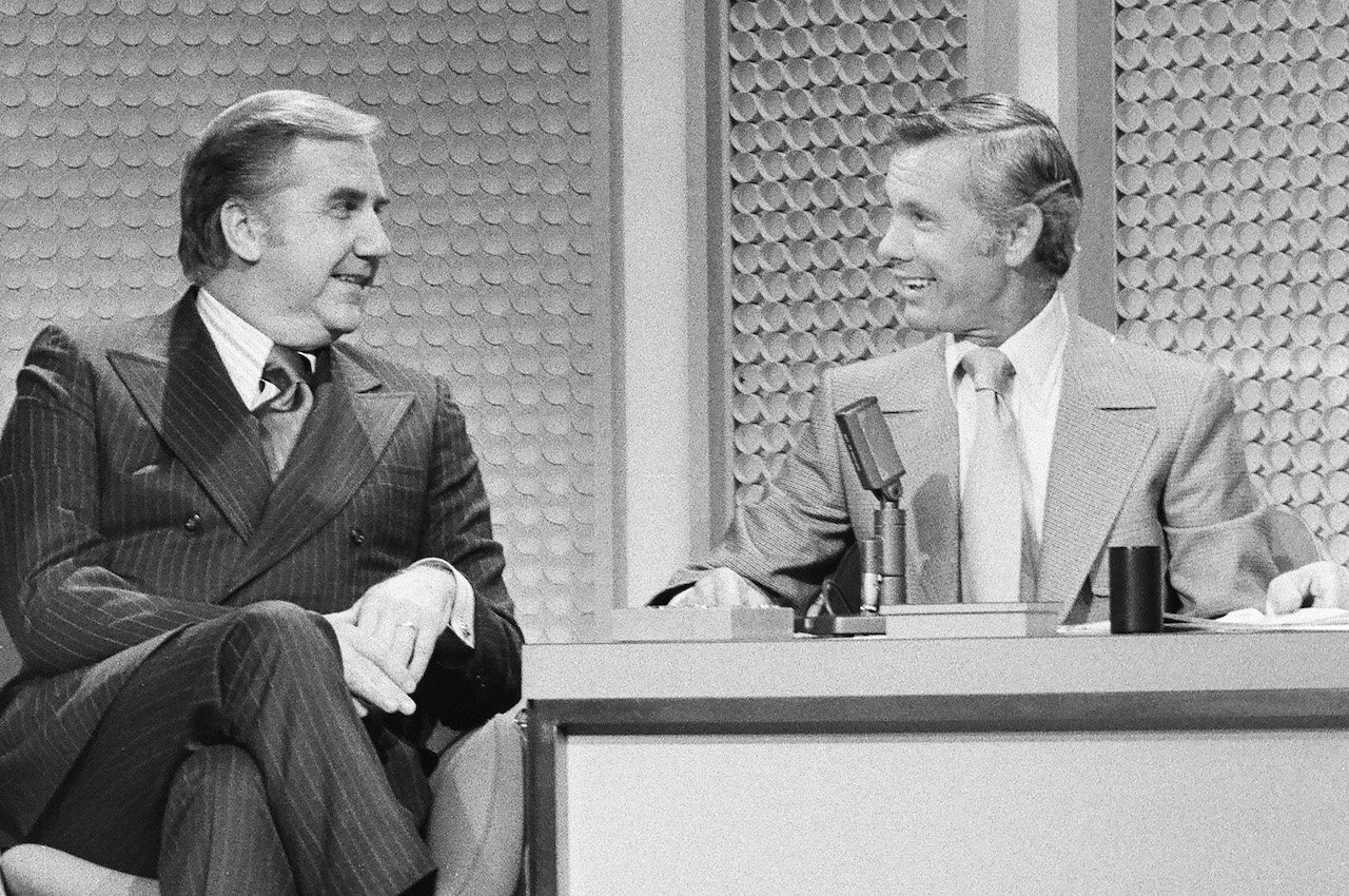 In black and white, Ed McMahon and Johnny Carson look at each other while sitting at 'The Tonight Show' desk in 1972