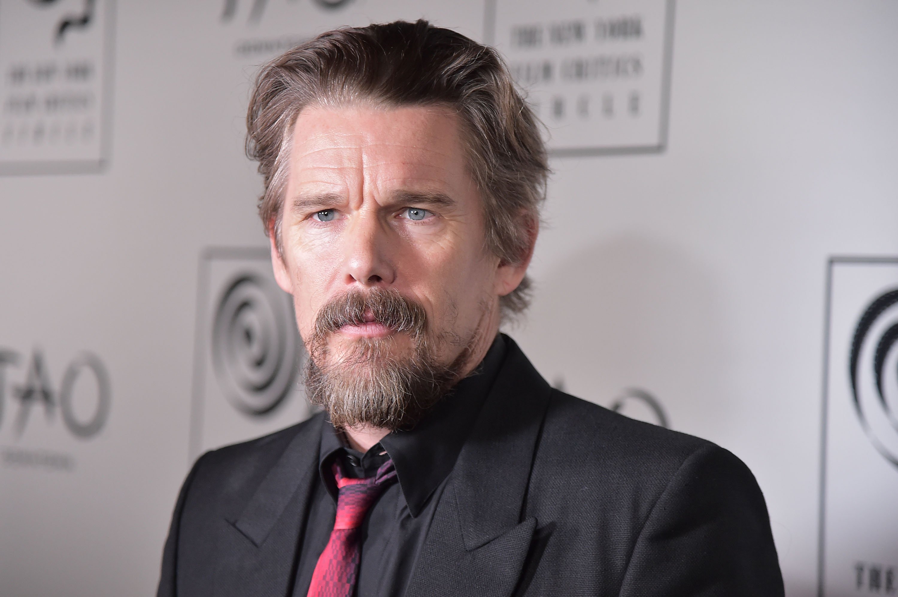 Ethan Hawke, who plays Arthur Harrow in 'Moon Knight,' wears a black suit over a black button-up shirt and red tie.