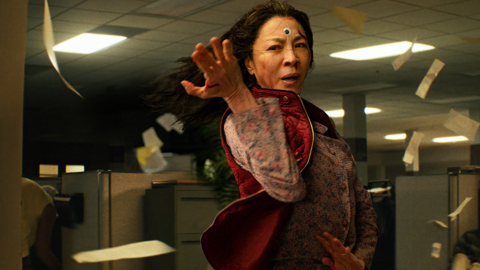 'Everything, Everywhere All at Once' Michelle Yeoh as Evelyn Wang in a fighting pose with a googly eye attached to her forehead and papers flying around