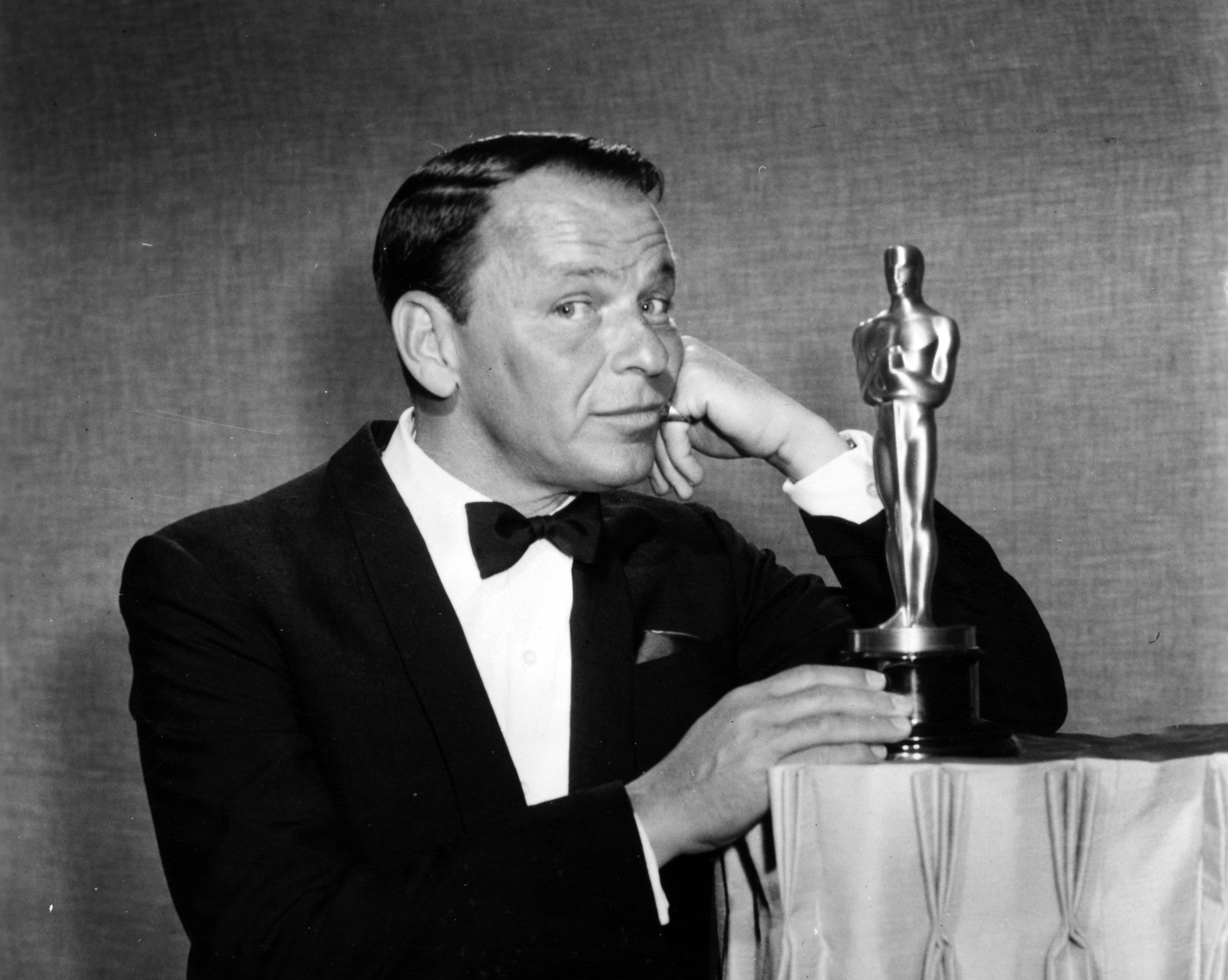 A black and white photo of Frank Sinatra in a tuxedo posing next to an Oscars statue. 