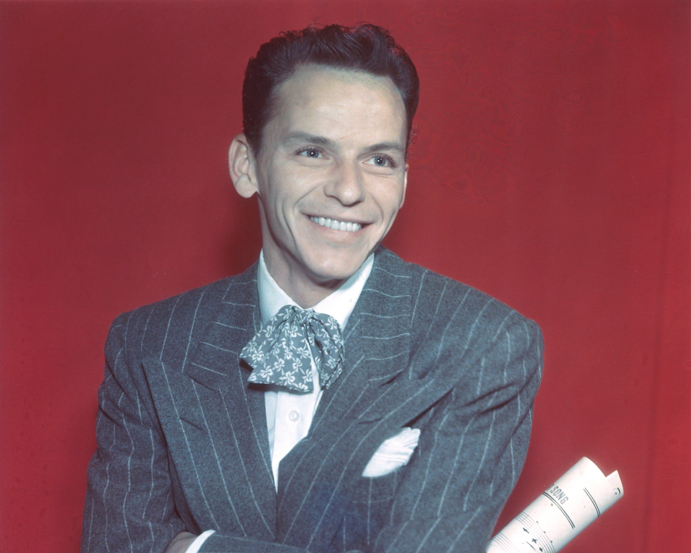 Frank Sinatra wears a suit and sits in front of a red background. 