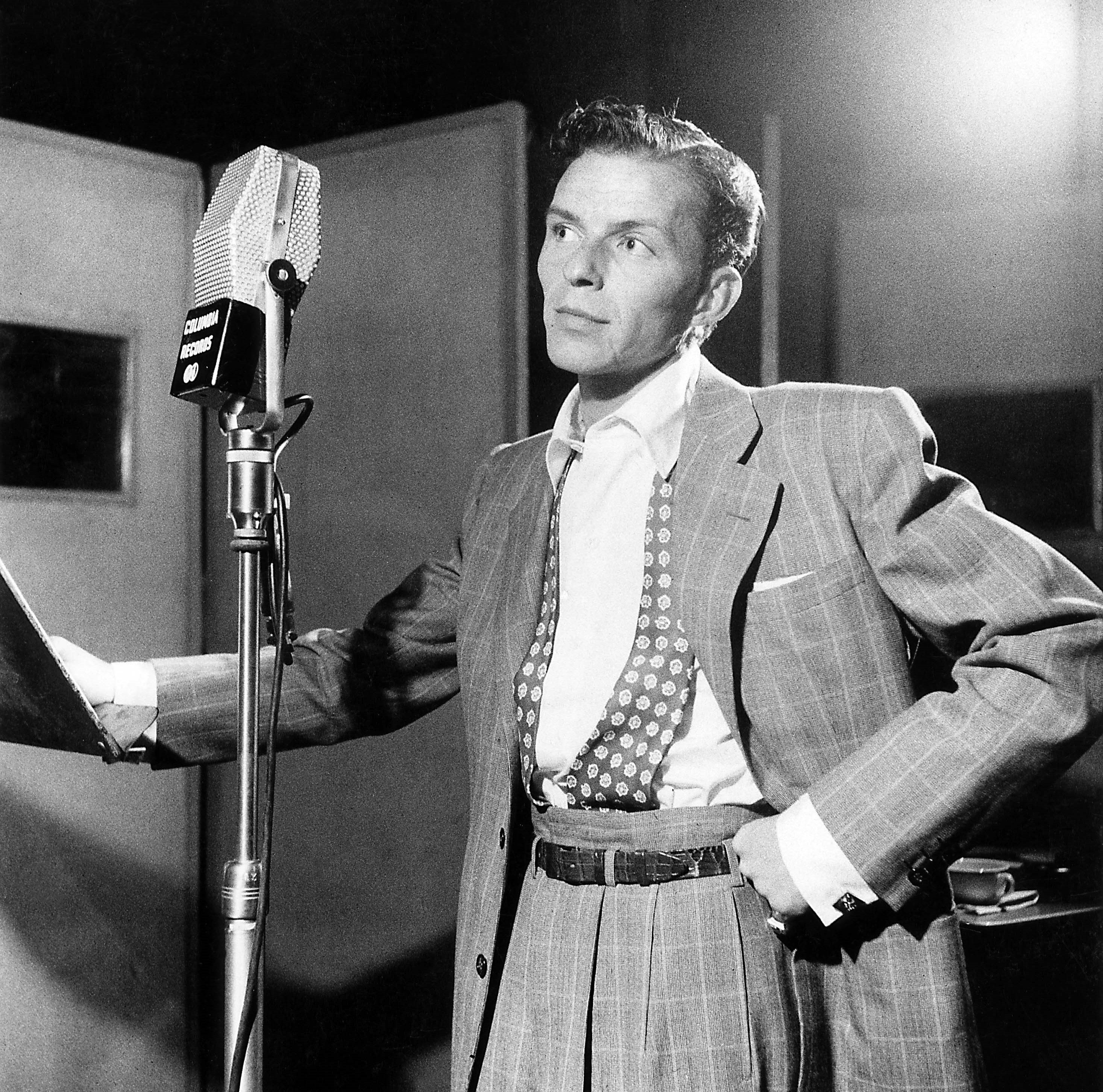 A black and white photo of Frank Sinatra standing in front of a microphone at a New York concert hall.