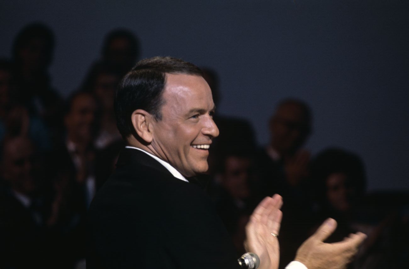 Frank Sinatra stands near a microphone and claps his hands.