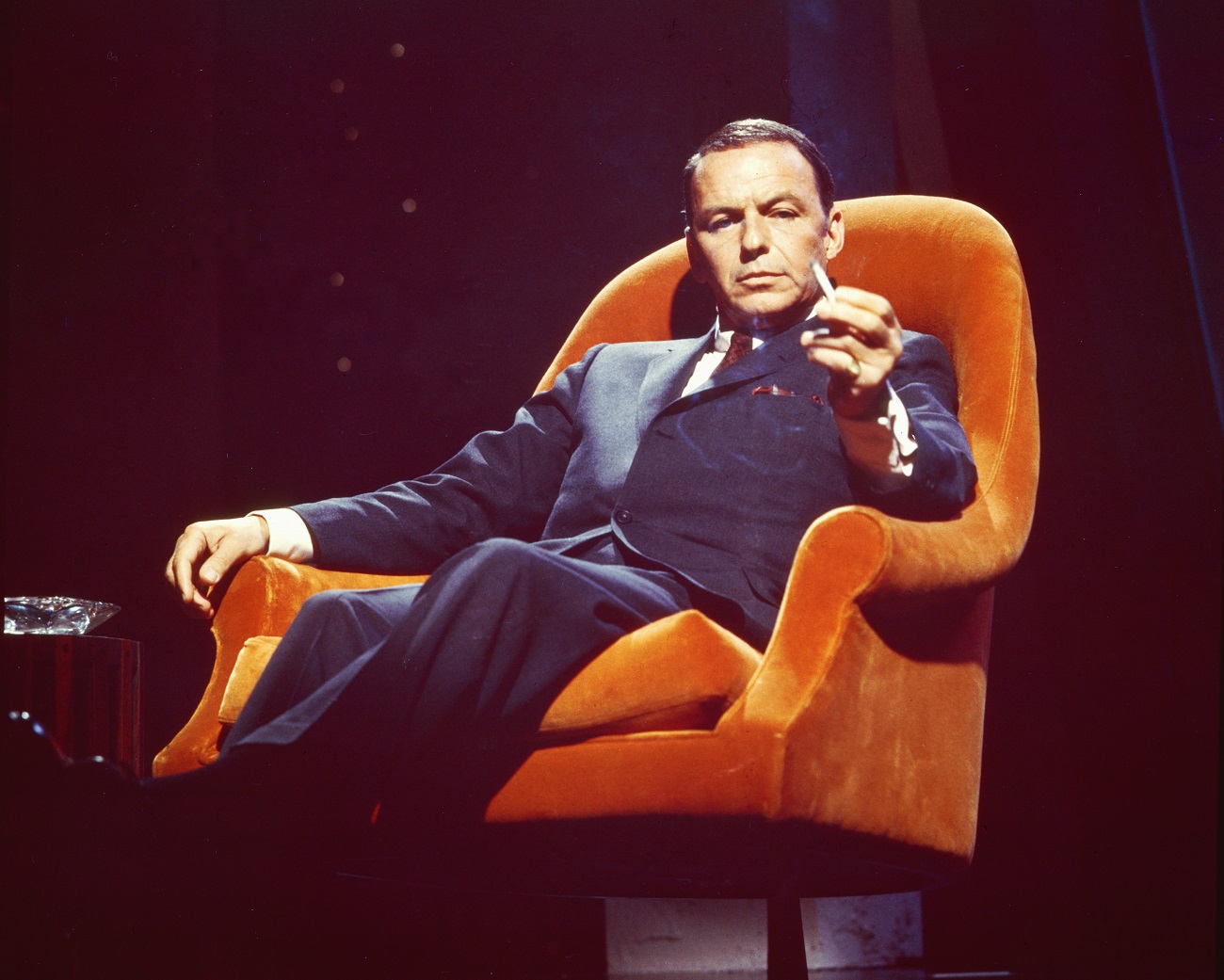 Frank Sinatra sits in an orange armchair and holds a cigarette.