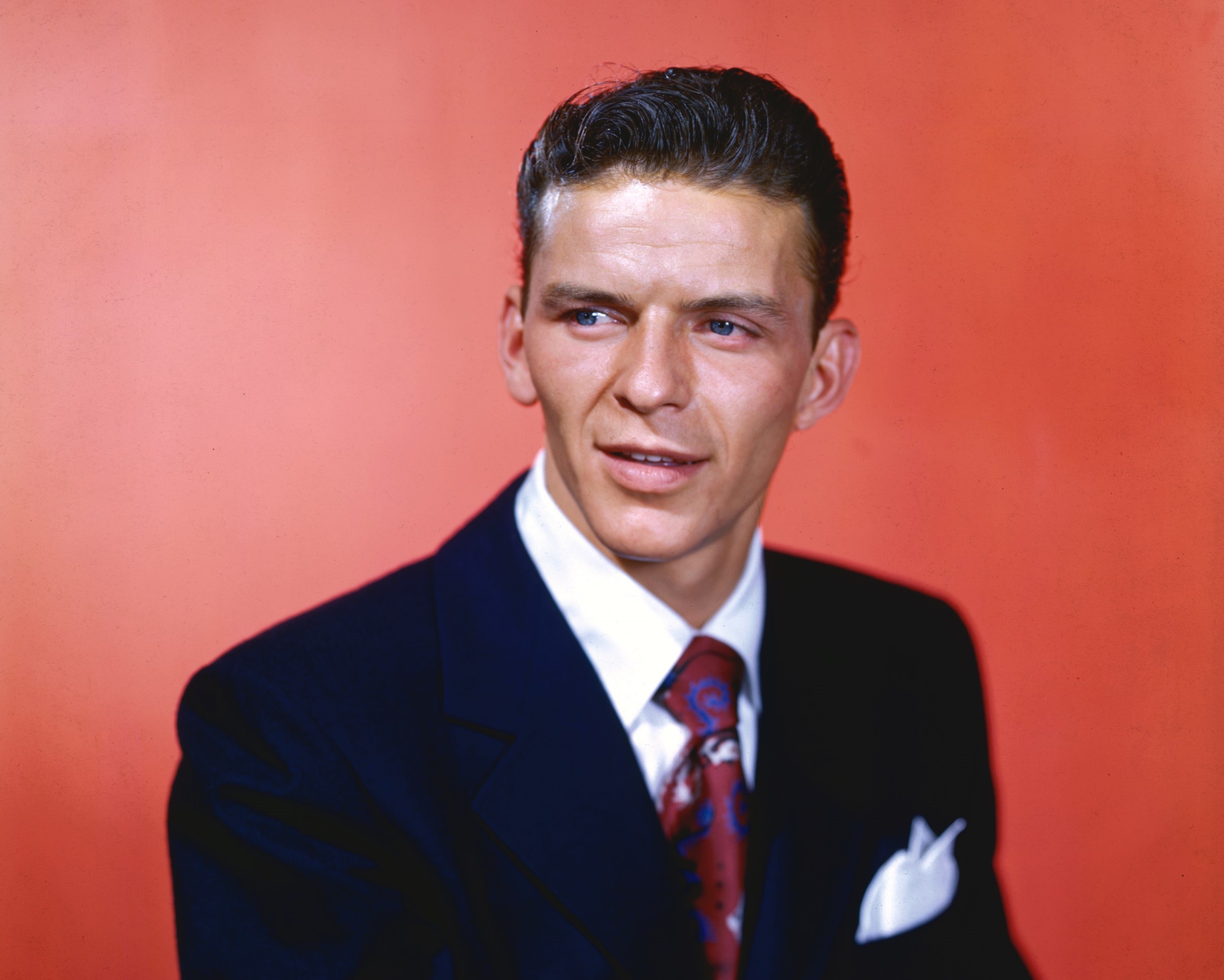 Frank Sinatra wears a suit and sits in front of a red background.