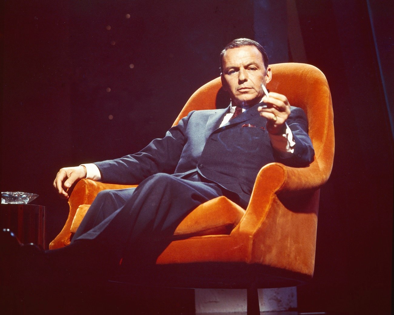Frank Sinatra wears a suit and sits in an orange armchair. He holds a cigarette.