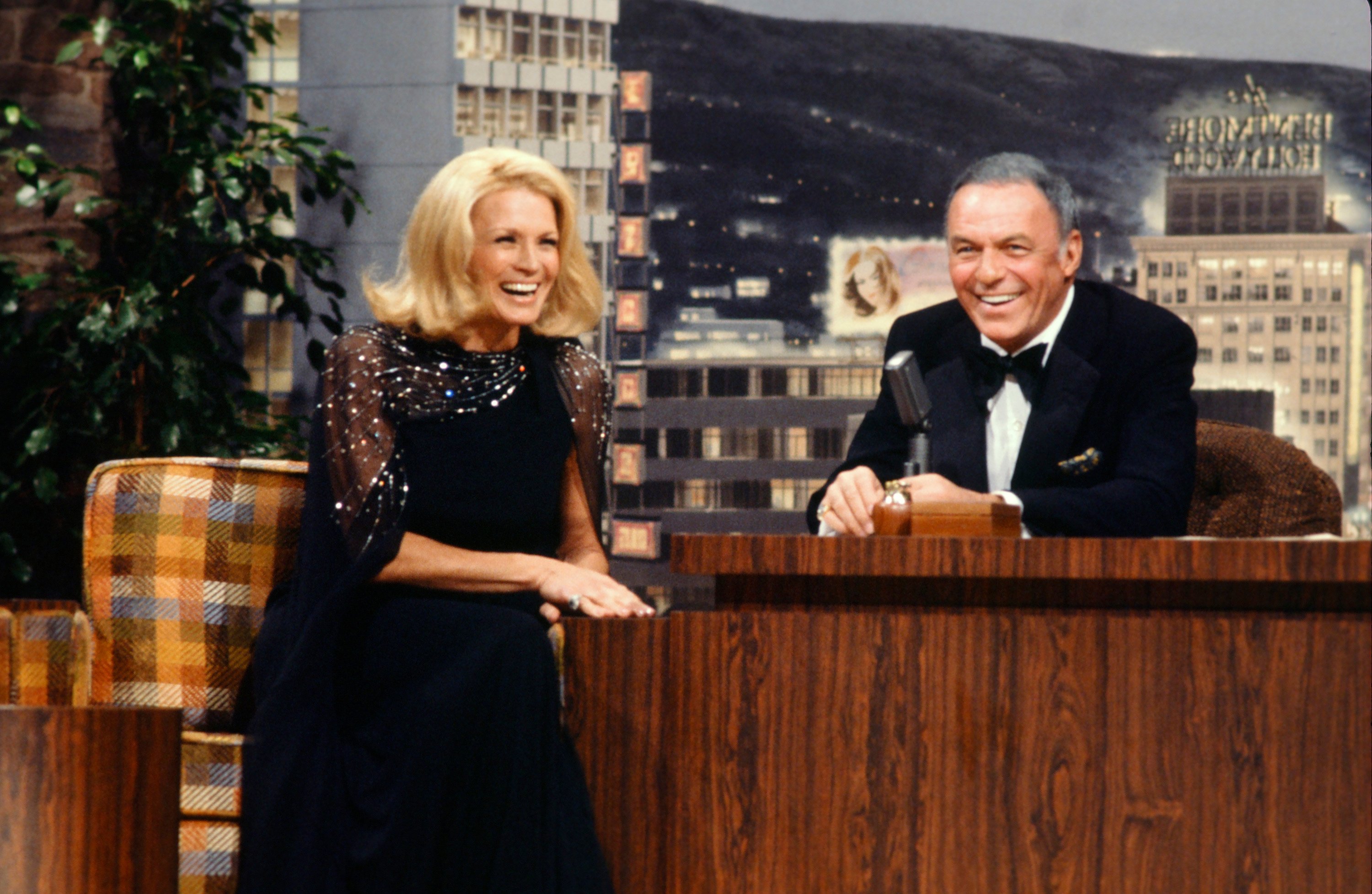 Angie Dickinson sits in a plaid arm chair and Frank Sinatra sits behind 'The Tonight Show' desk as a guest host.