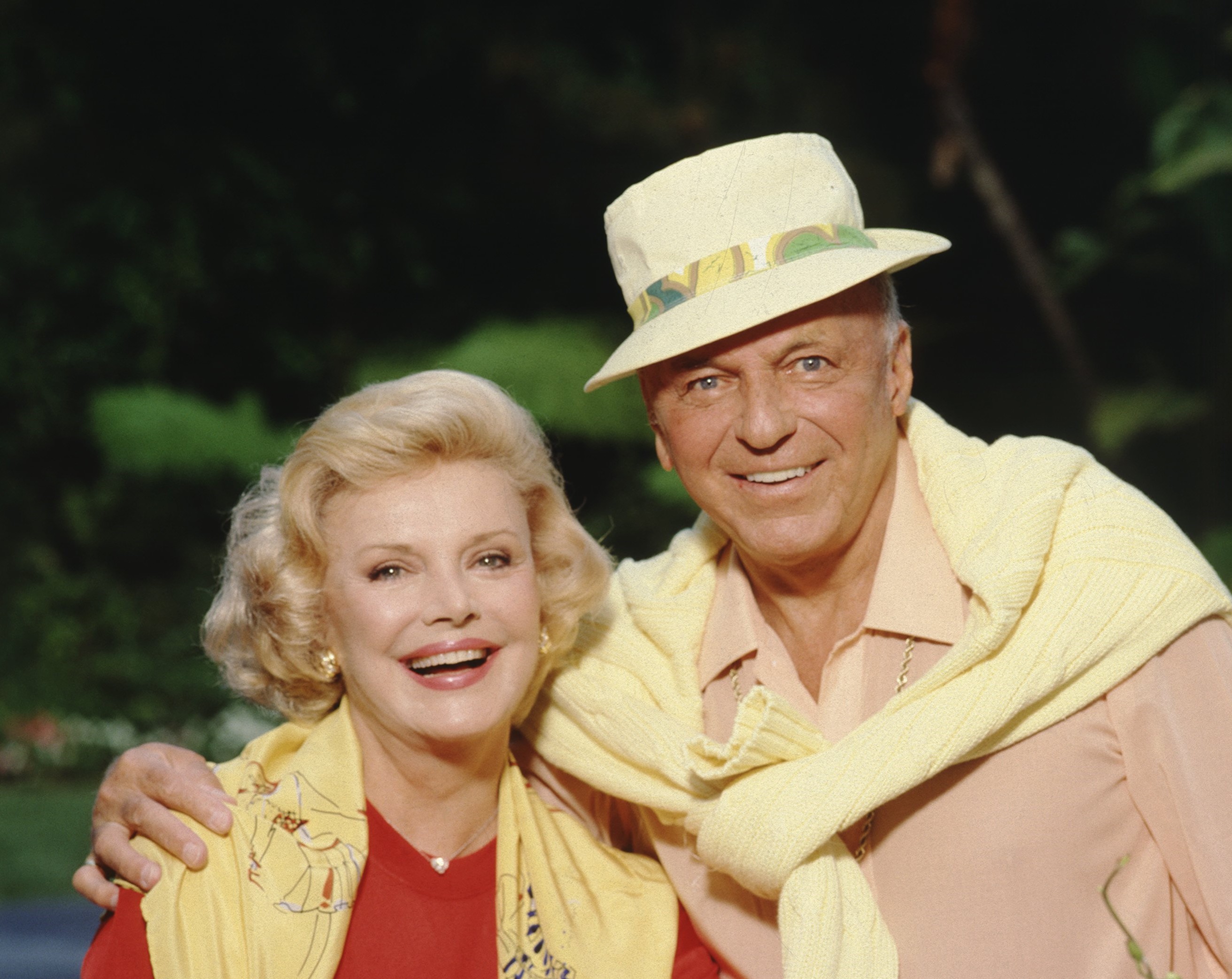Frank Sinatra has his hand around his wife Barbara Sinatra's shoulders. They both have yellow sweaters hanging over their shoulders.