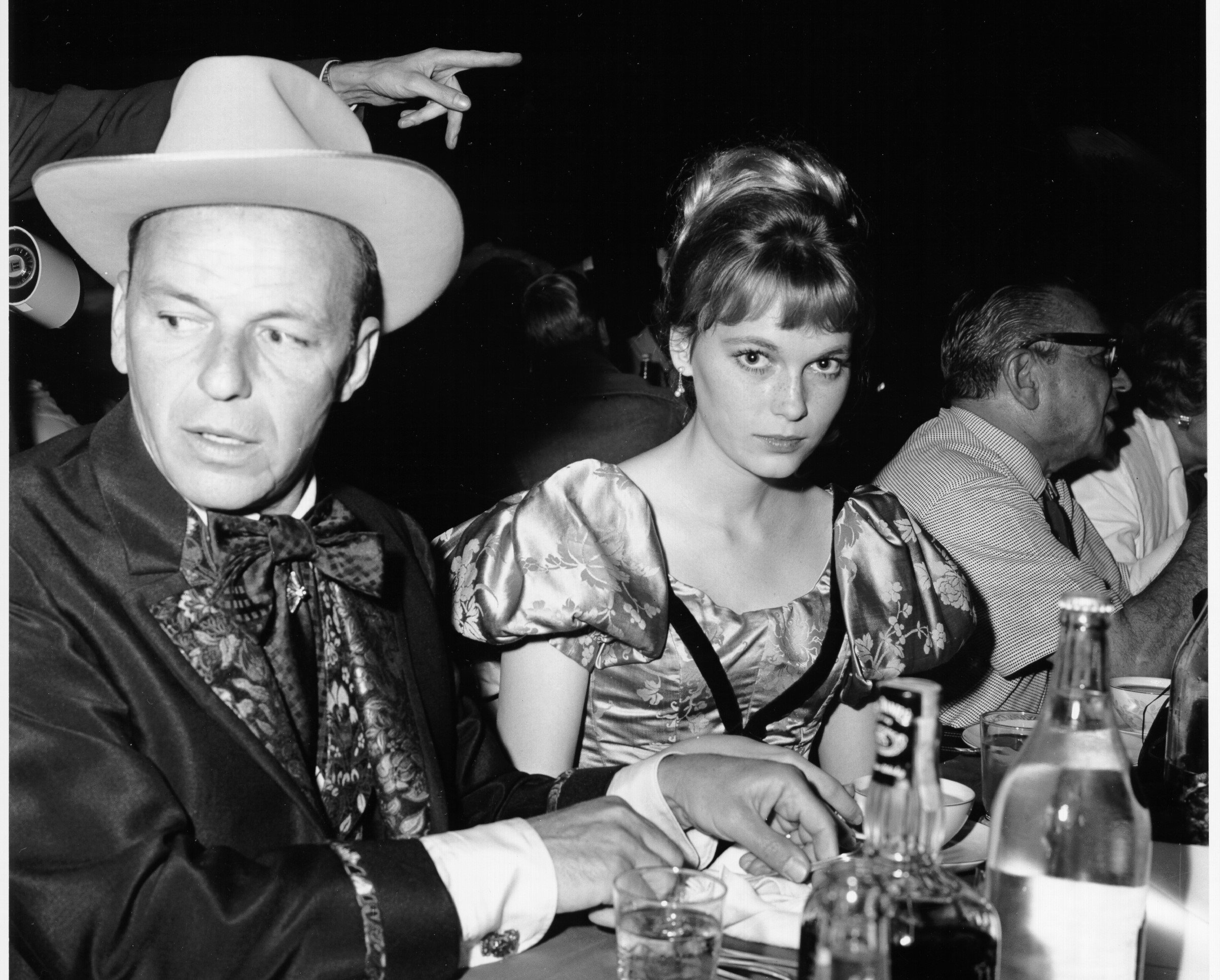 A black and white photo of Frank Sinatra and Mia Farrow sitting at a table. He wears a hat and she wears a dress with poofed sleeves.