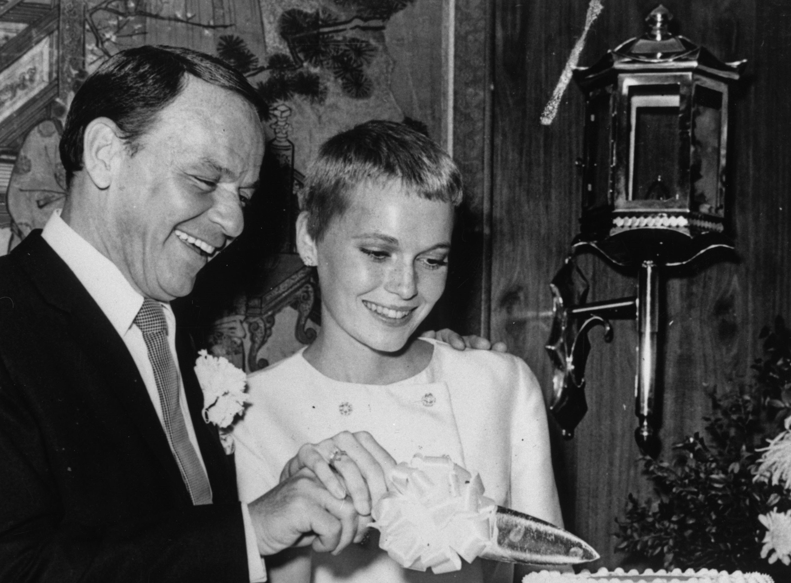 A black and white photo of Frank Sinatra and Mia Farrow cutting into a cake on their wedding day.