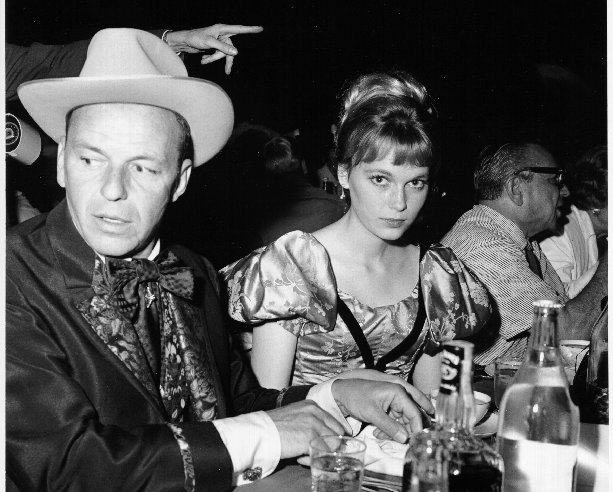 A black and white photo of Frank Sinatra and Mia Farrow sitting at a table. He wears a hat and she wears a dress with puffed sleeves.