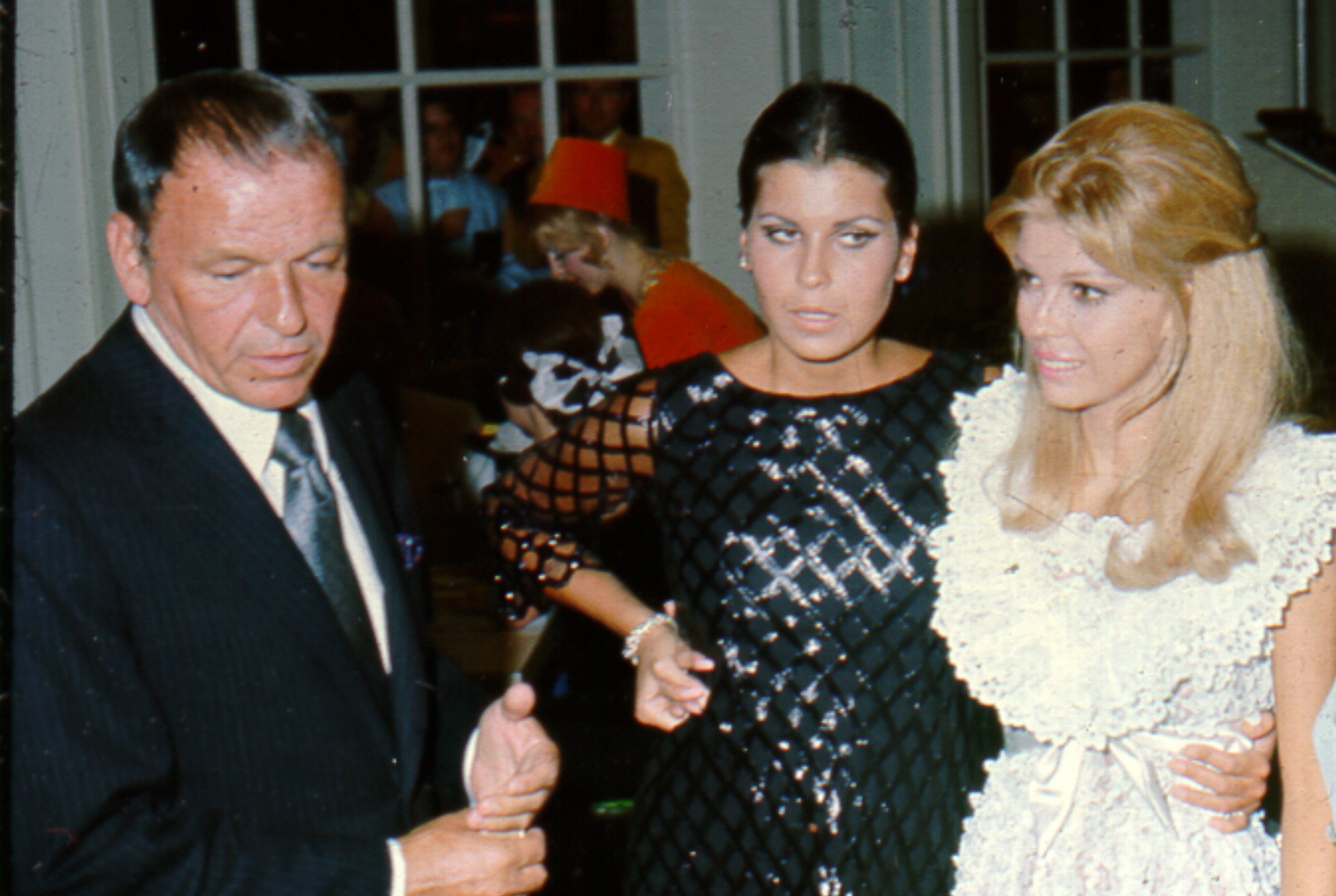 Frank Sinatra wears a suit, Tina Sinatra wears a black dress, and Nancy Sinatra wears a white dress at a party.