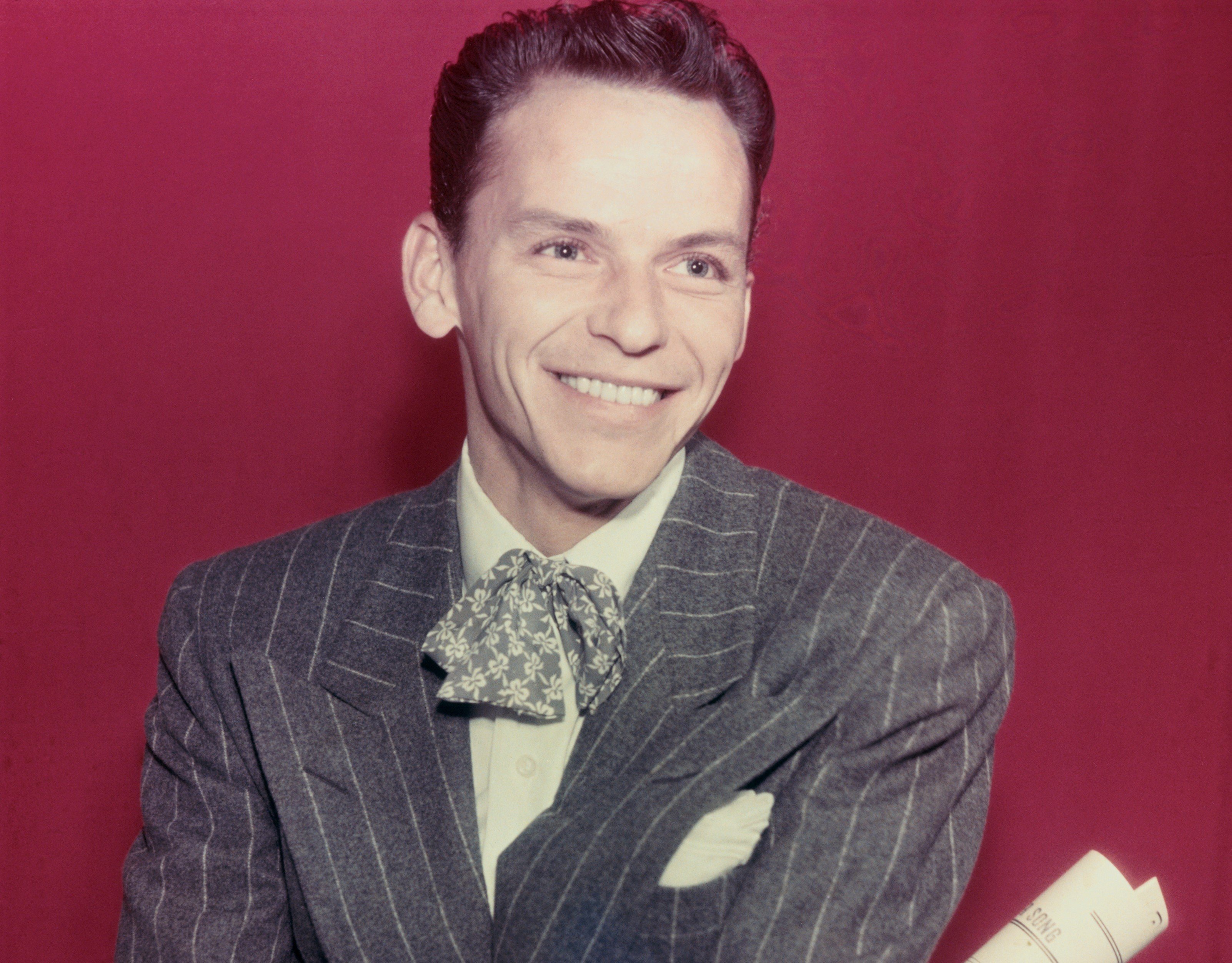 Frank Sinatra wears a gray suit and holds a rolled up paper. He sits against a maroon background.