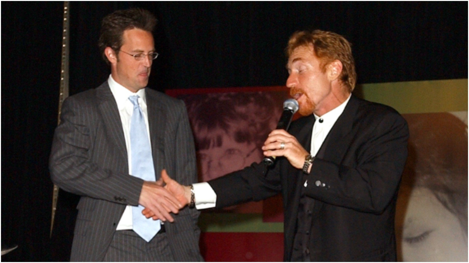 Matthew Perry and Danny Bonaduce shake hands on stage as Bonaduce holds a microphone 