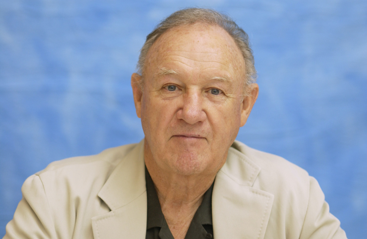 Gene Hackman headshot wearing a tan blazer against a blue background