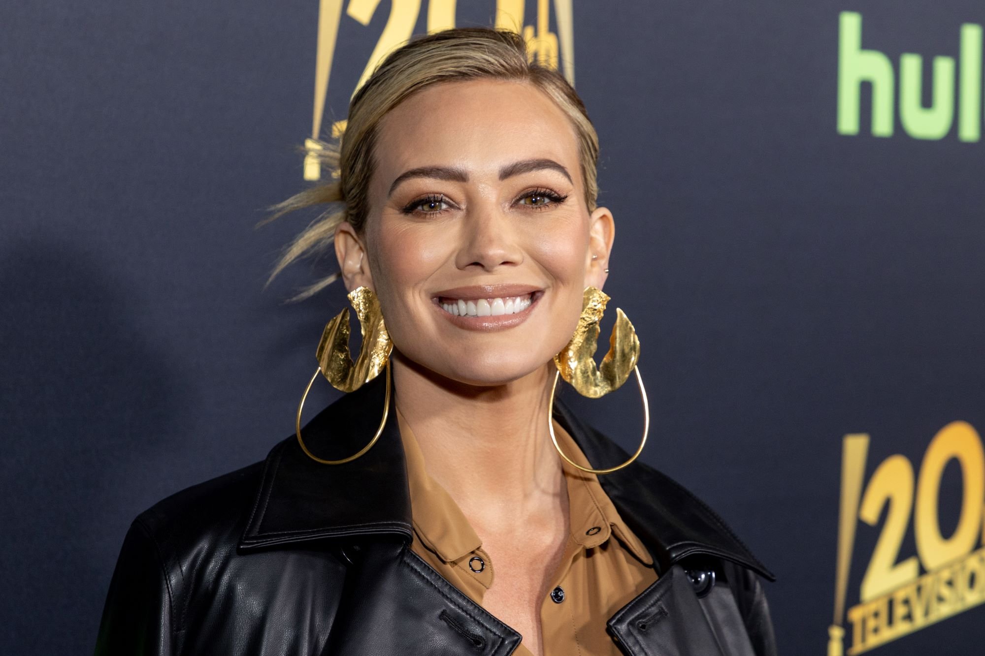Hilary Duff, who stars in 'How I Met Your Father' Season 2, wears a black leather jacket over a light brown button-up shirt and gold dangly earrings.