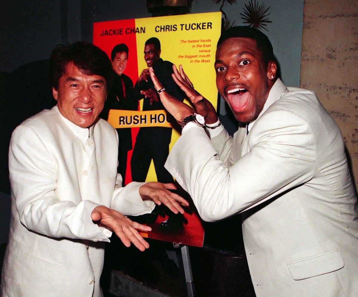 A smiling Jackie Chan and Chris Tucker wear white and high five in front of the ‘Rush Hour’ poster