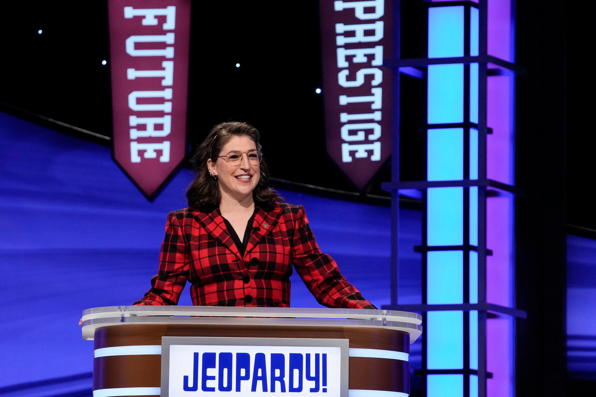 'Jeopardy!' host Mayim Bialik stands at the podium wearing a red blazer