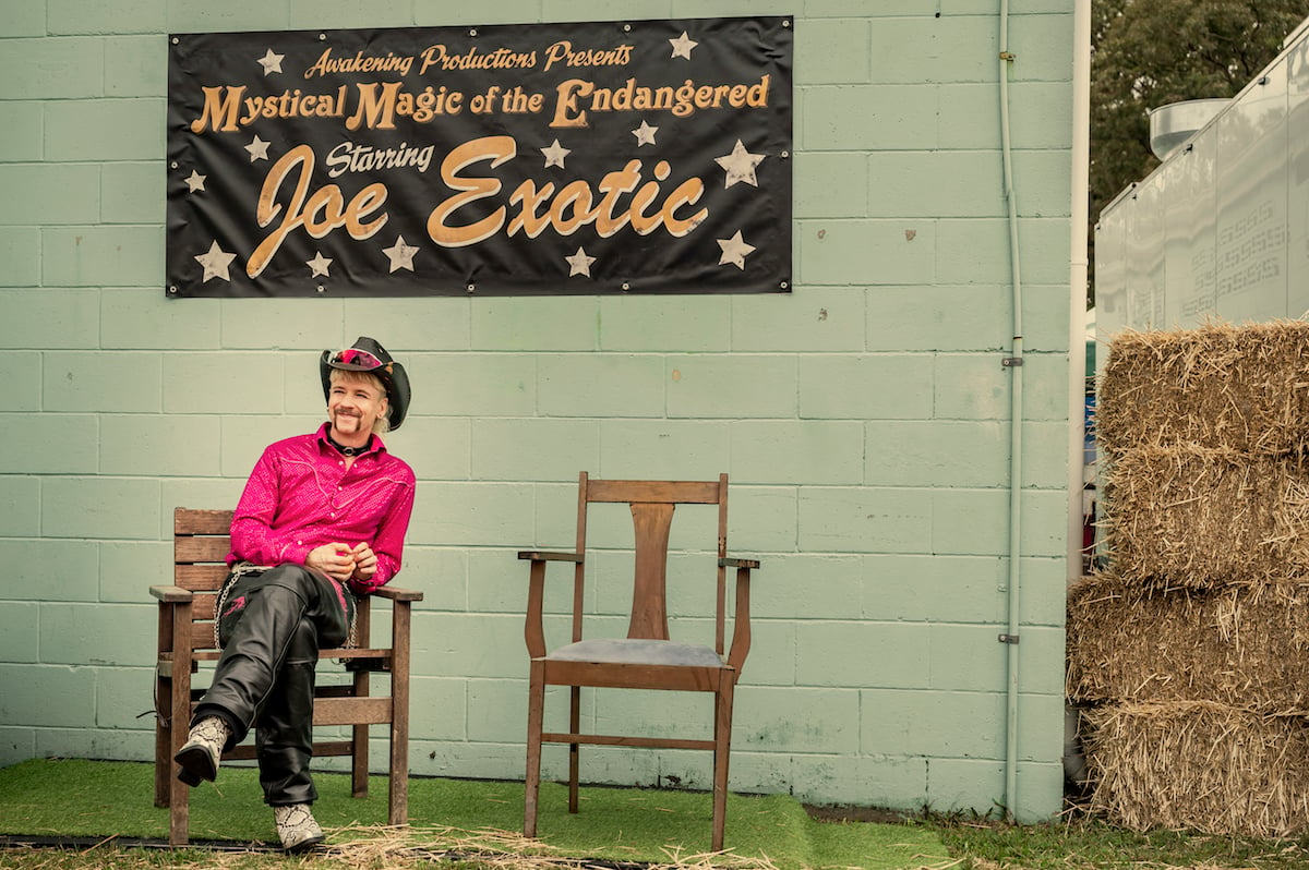 John Cameron Mitchell as Joe Exotic, wearing a pink shirt and sitting in front of a sign for his show, in 'Joe vs Carole' 