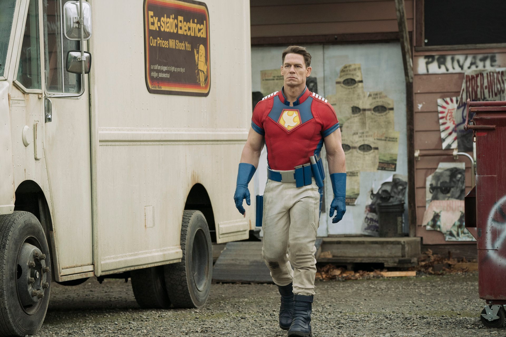 John Cena, who will return as Christopher Smith in ‘Peacemaker’ Season 2, in costume walking next to a large white truck, and he's wearing beige pants, a red shirt, and blue gloves.