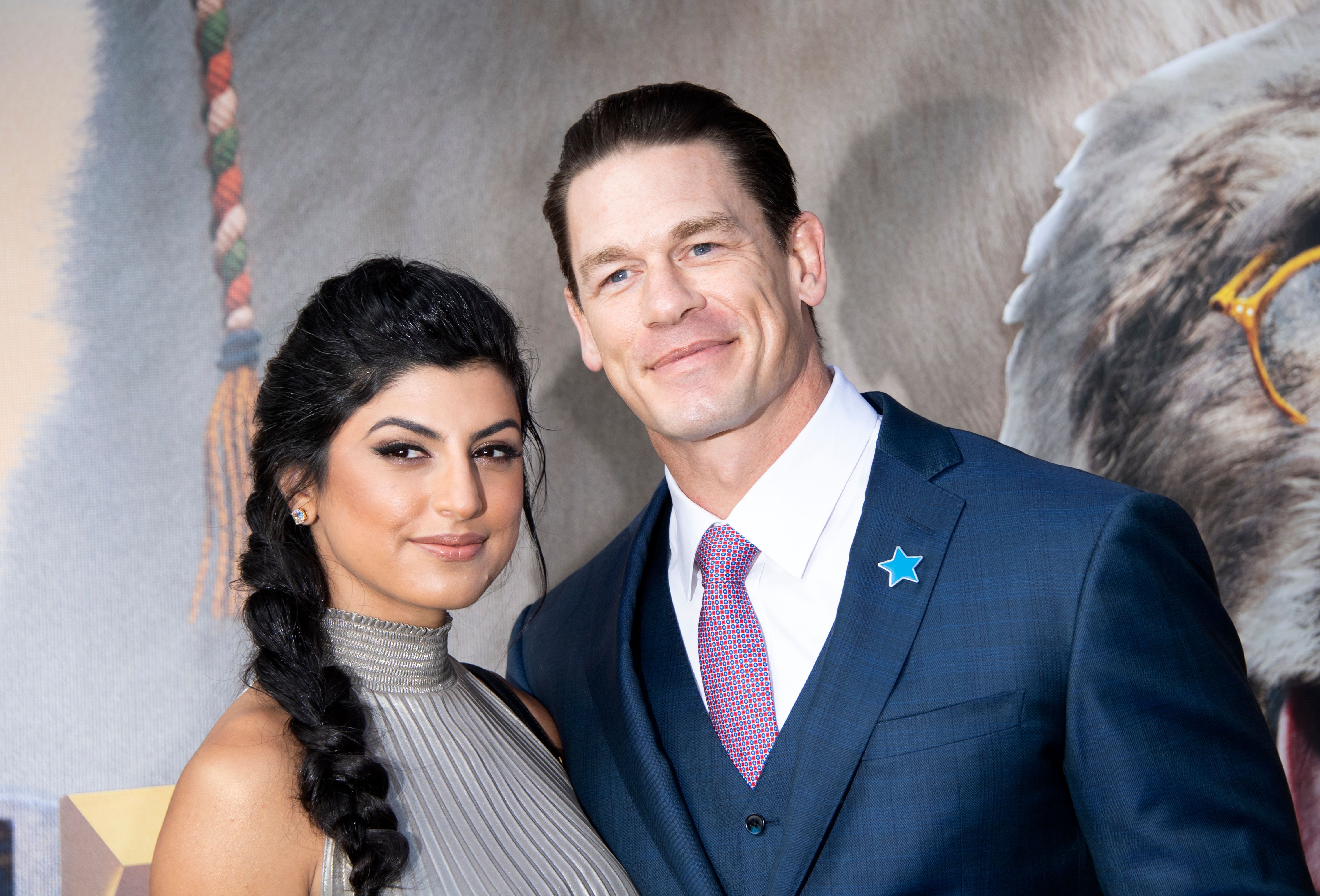 Wrestler and actor John Cena wearing a navy blue suit poses with his wife Shay Shariatzadeh on the red carpet.