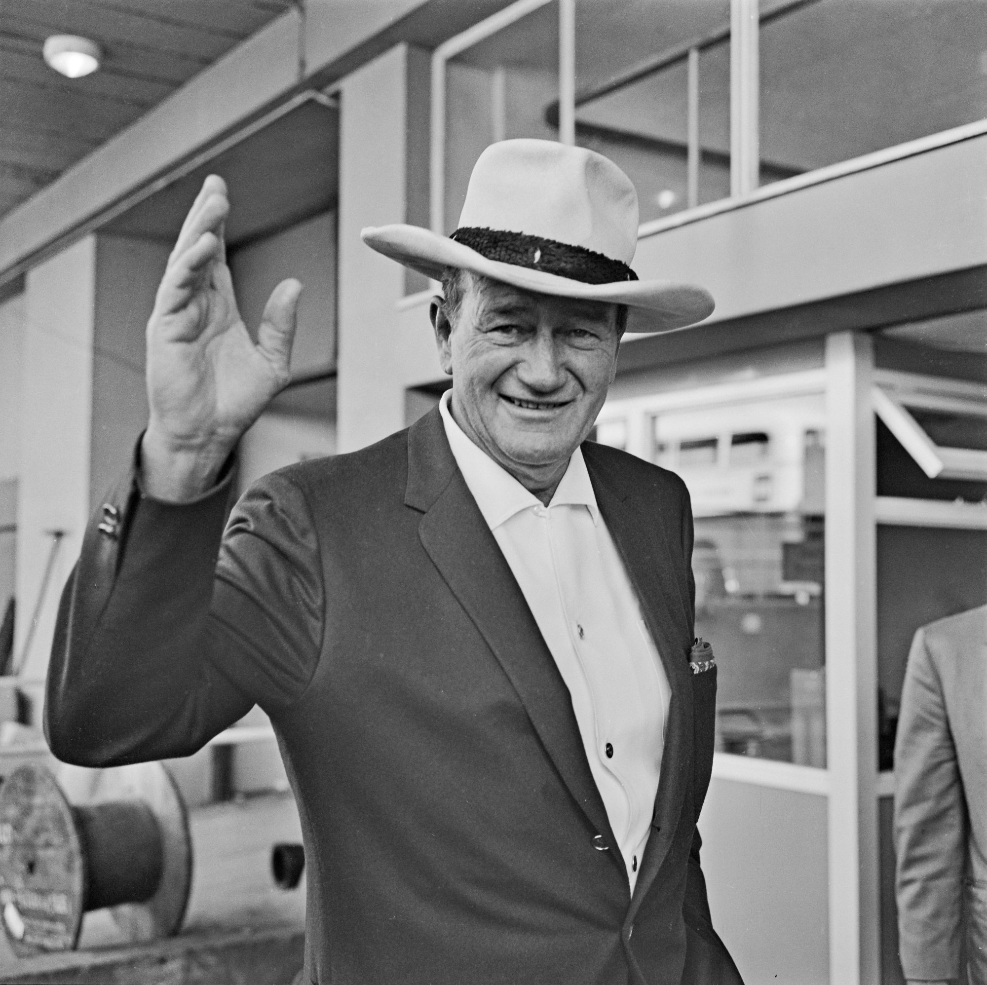 John Wayne holding a hand up waving wearing a suit and hat