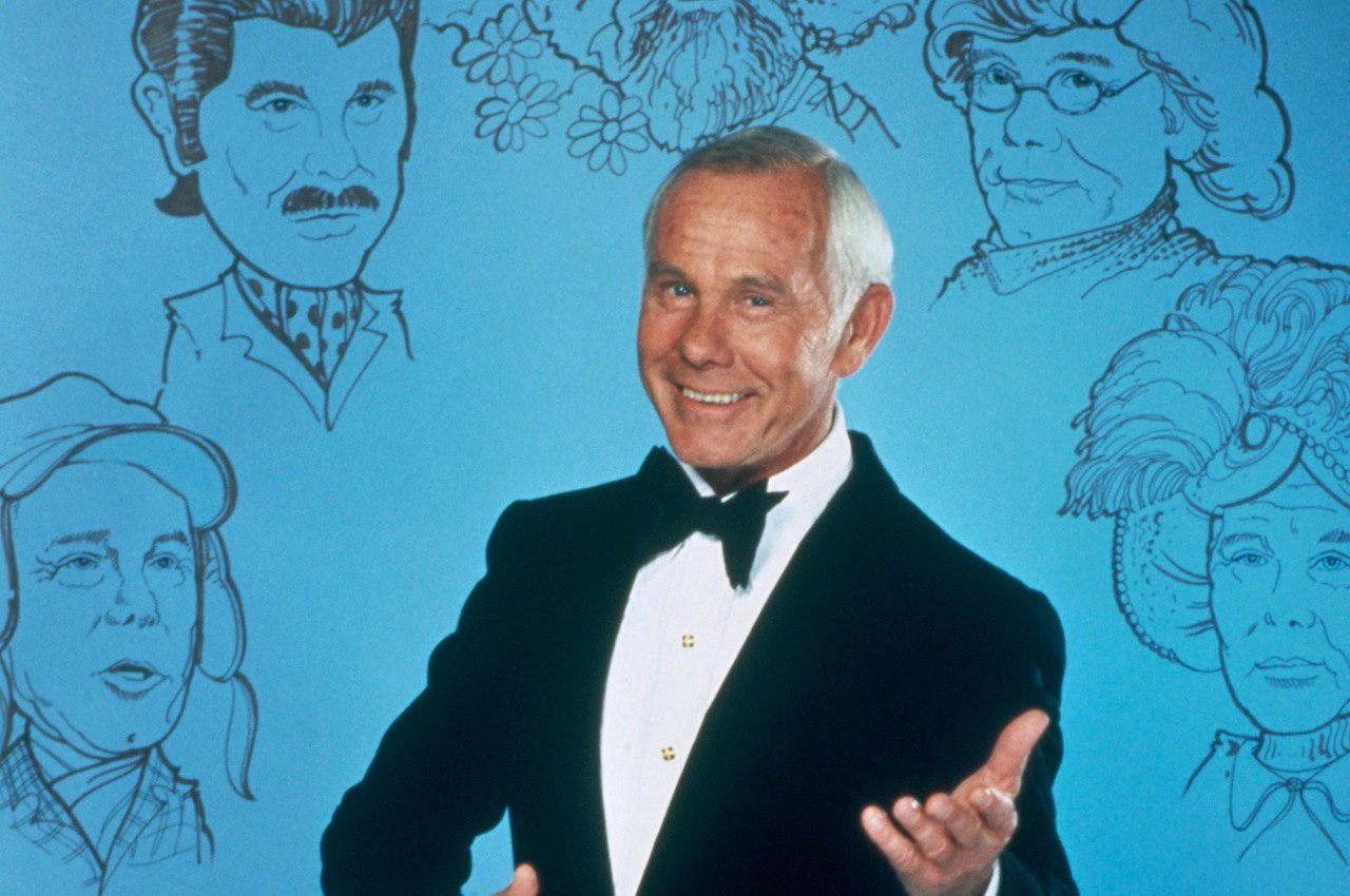 Johnny Carson stands in front of a blue background decorated with drawings of some of his famous sketch characters from 'The Tonight Show.'