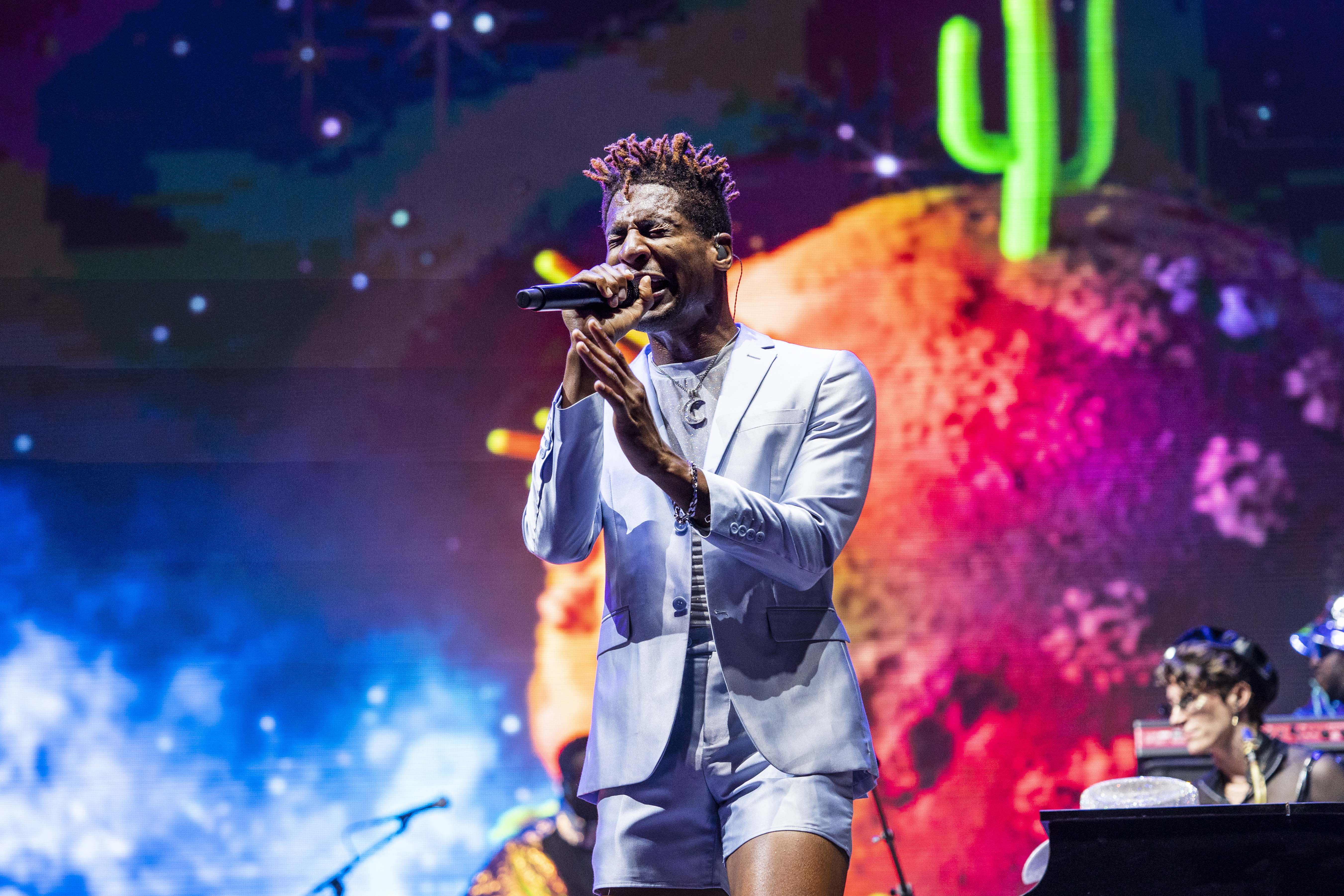 Jon Batiste sings into a microphone while standing on a colorful stage.