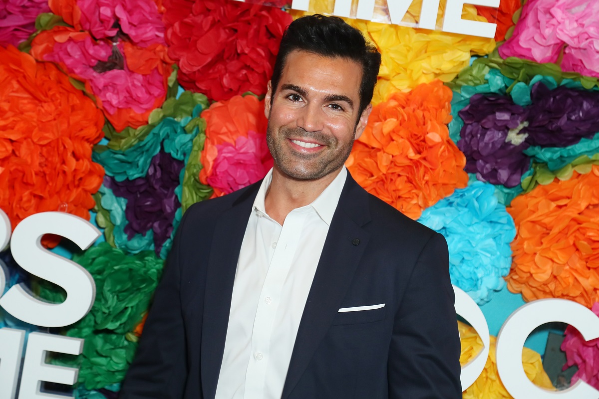 'The Young and the Restless' actor Jordi Vilasuso wearing a navy blue suit and white shirt; standing in front of a floral hedge.
