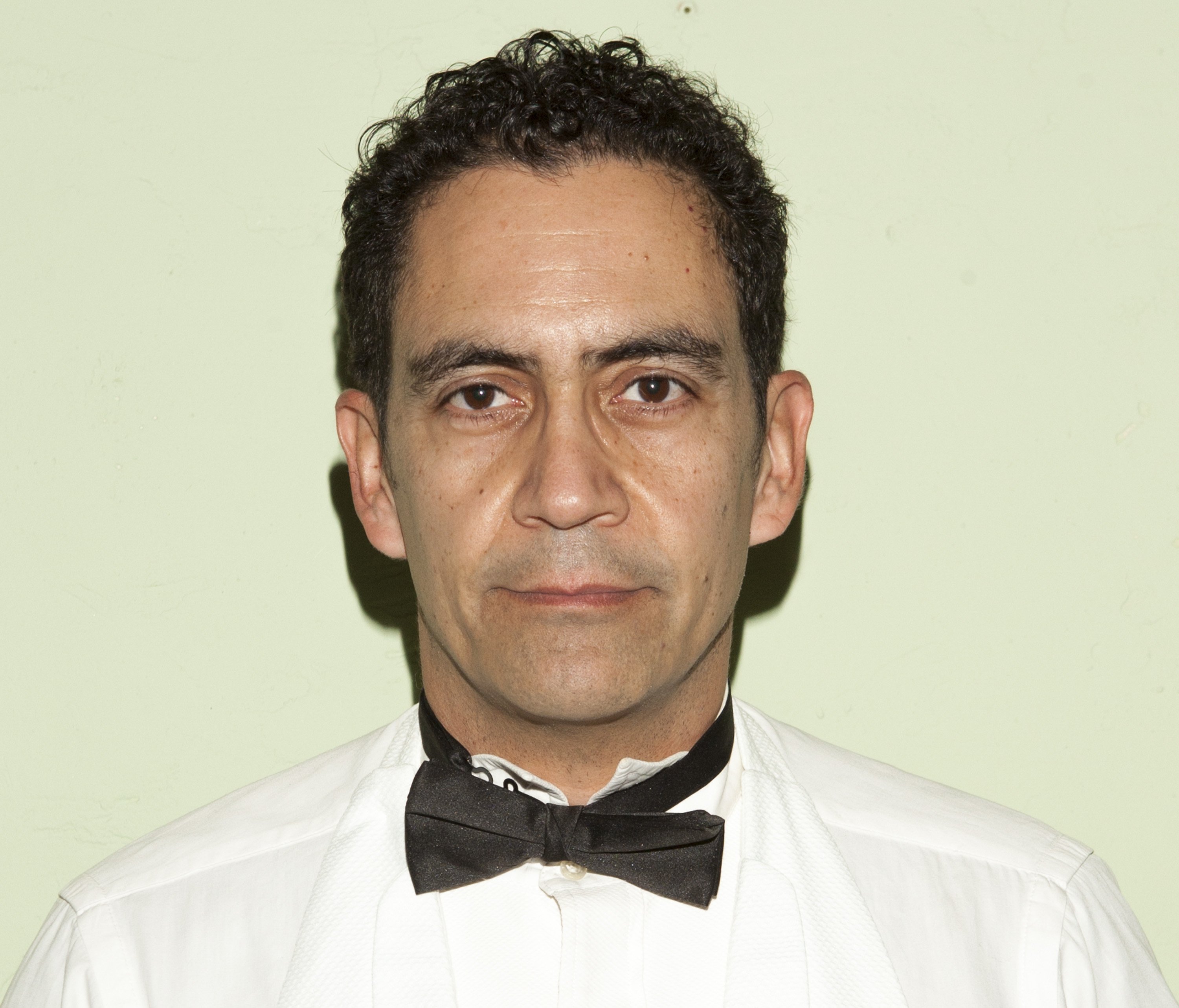 Jose Zuniga who plays Javier Escano on Chicago P.D. Season 9 wearing a white button-up shirt and a black bow tie.