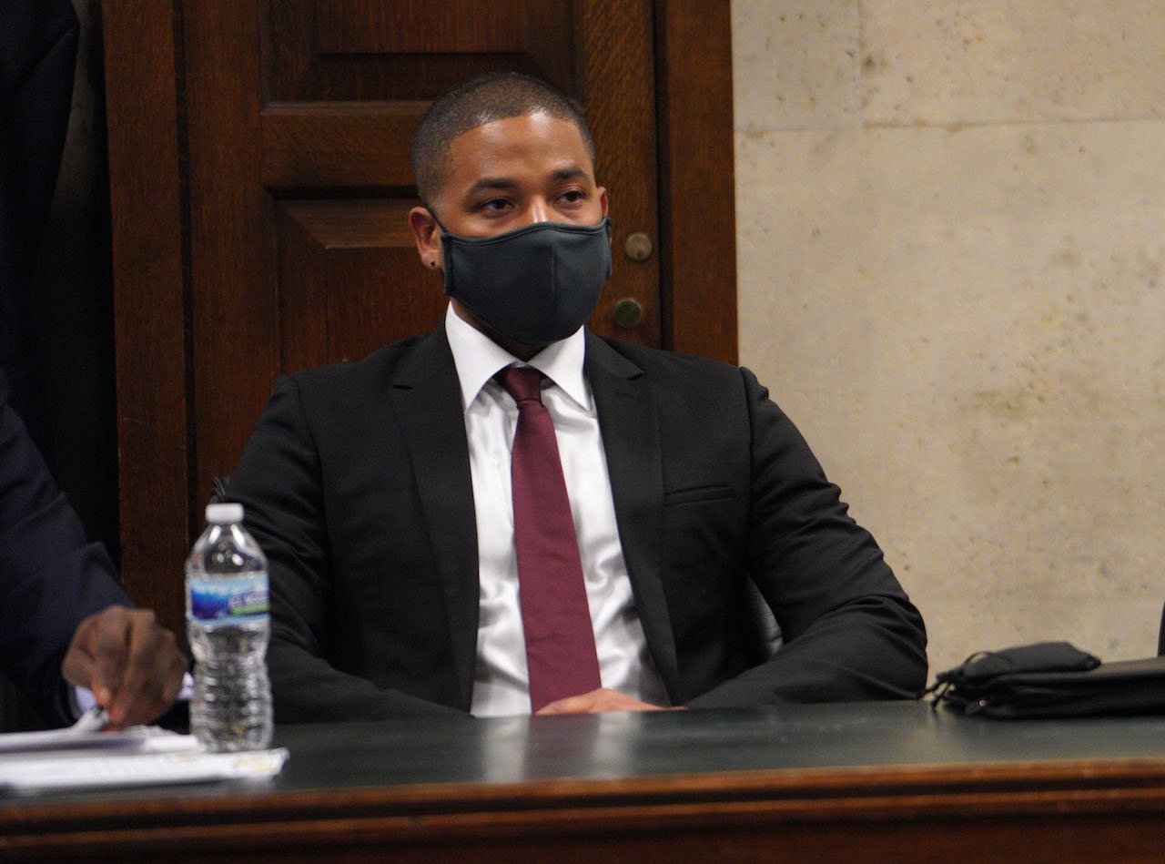 Jussie Smollett in a suit and tie wearing a black face mask and seated a t a table at his sentencing