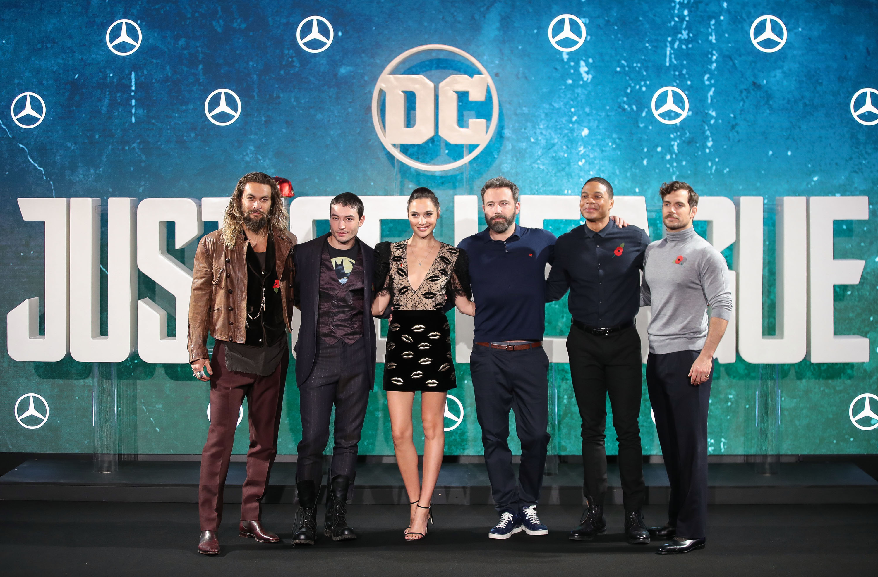 Jason Momoa, Ezra Miller, Gal Gadot, Ben Affleck, Ray Fisher, and Henry Cavill, the cast of 'Zack Snyder's Justice League,' which won Oscars Cheer Moment, pose for photos. Momoa wears a brown jacket over a black vest, gray shirt, and maroon pants. Miller wears a black suit over a dark red vest, and black pants. Gadot wears a black and tan dress with kisses on it. Affleck wears a blue polo shirt and black pants. Fisher wears a black button-up shirt and black pants. Cavill wears a light gray turtleneck and black pants.