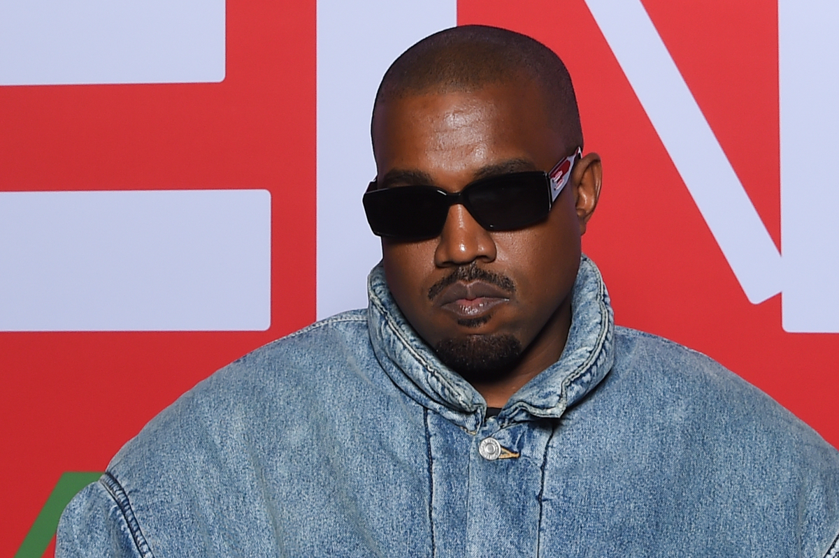 Kanye West in a denim jacket and black sunglasses poses with a stern face.
