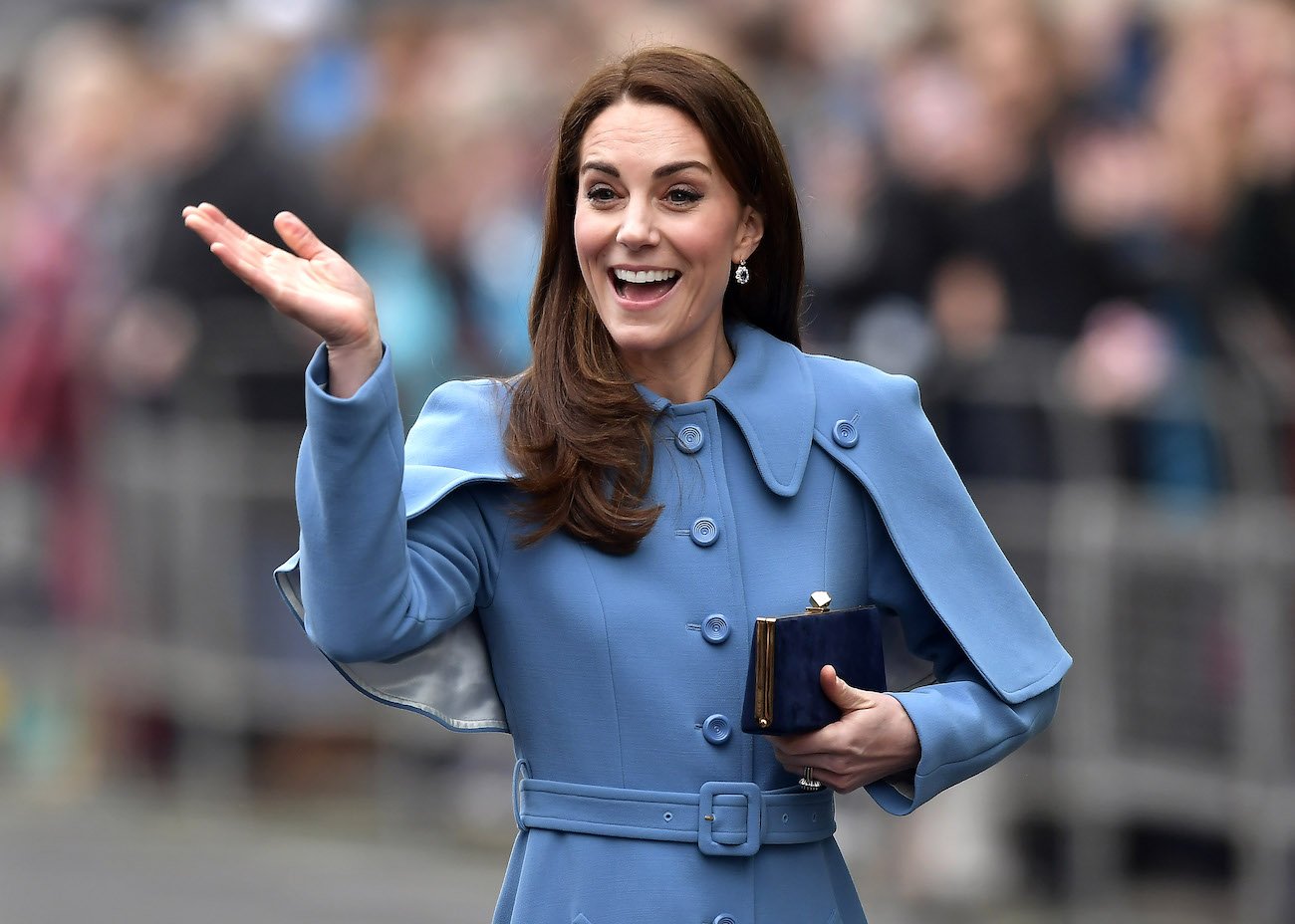 Kate Middleton waving with her right hand while wearing a blue coat