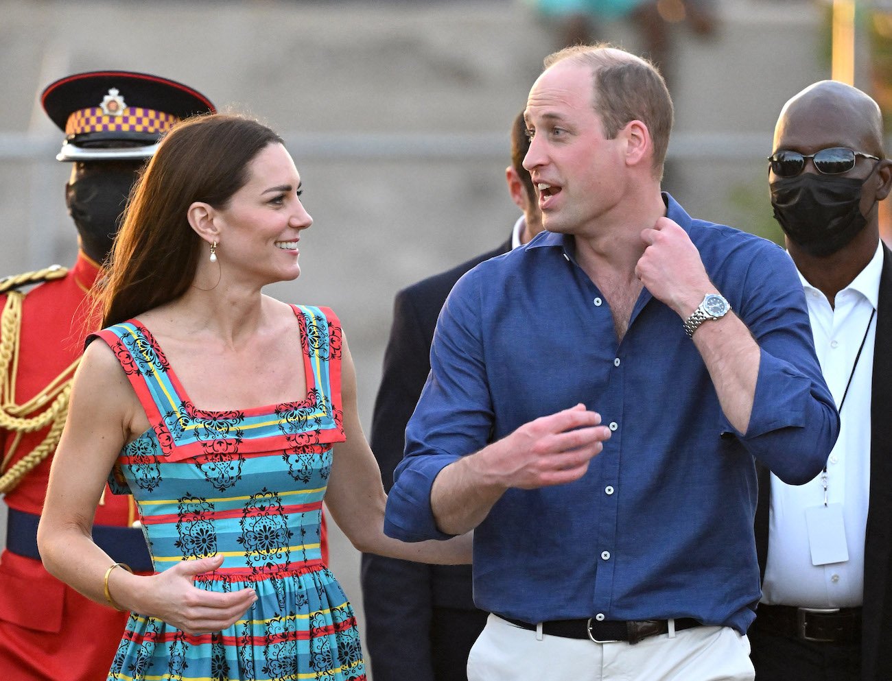Kate Middleton wearing a colorful sleeveless dress and walking beside Prince William, who is wearing a blue button down shirt with white pants