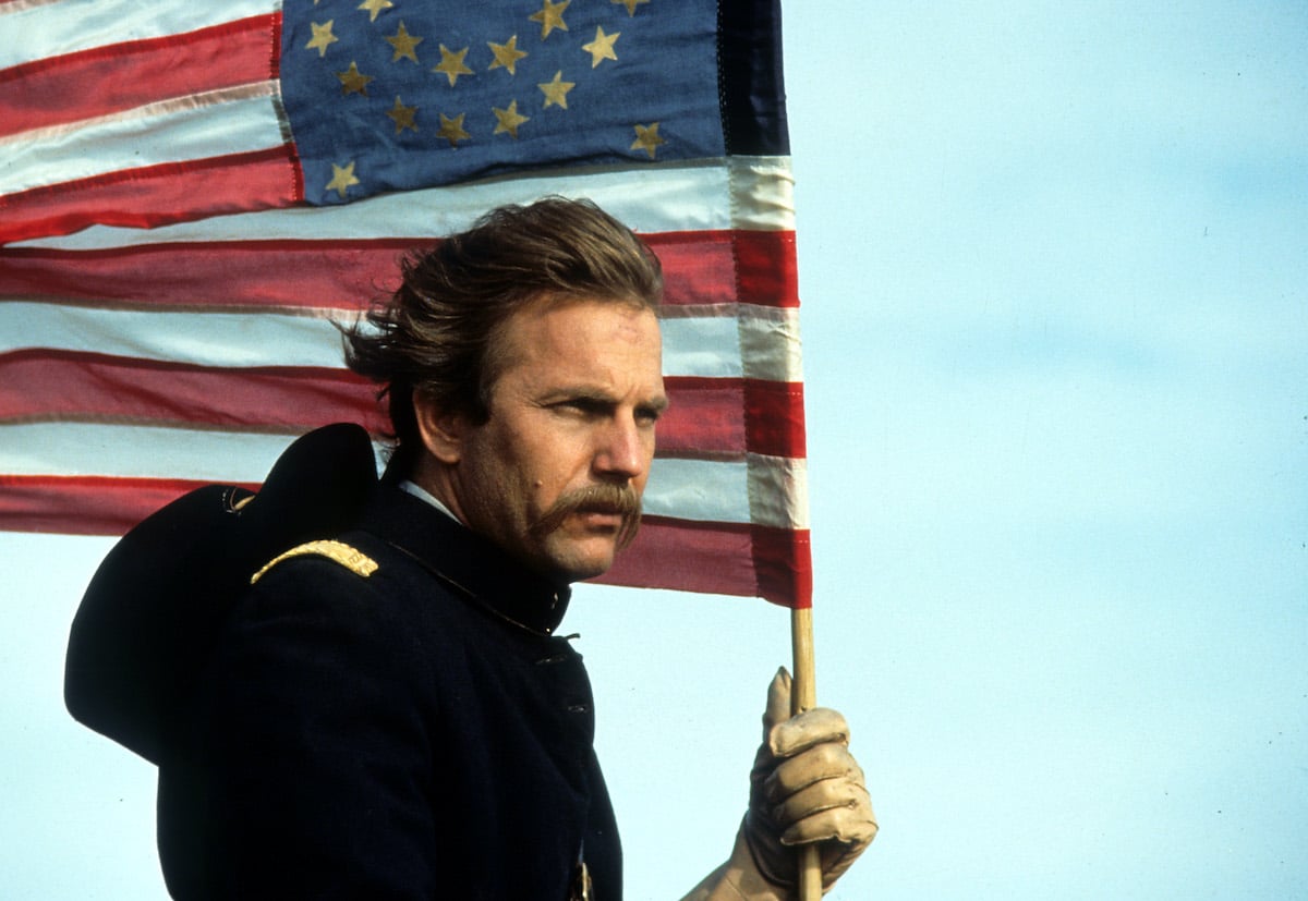 Kevin Costner holds an American flag and looks off into the distance
