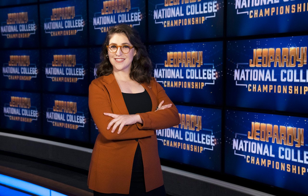 Jeopardy host Mayim Bialik in front of the Jeopardy! National College Championship logo